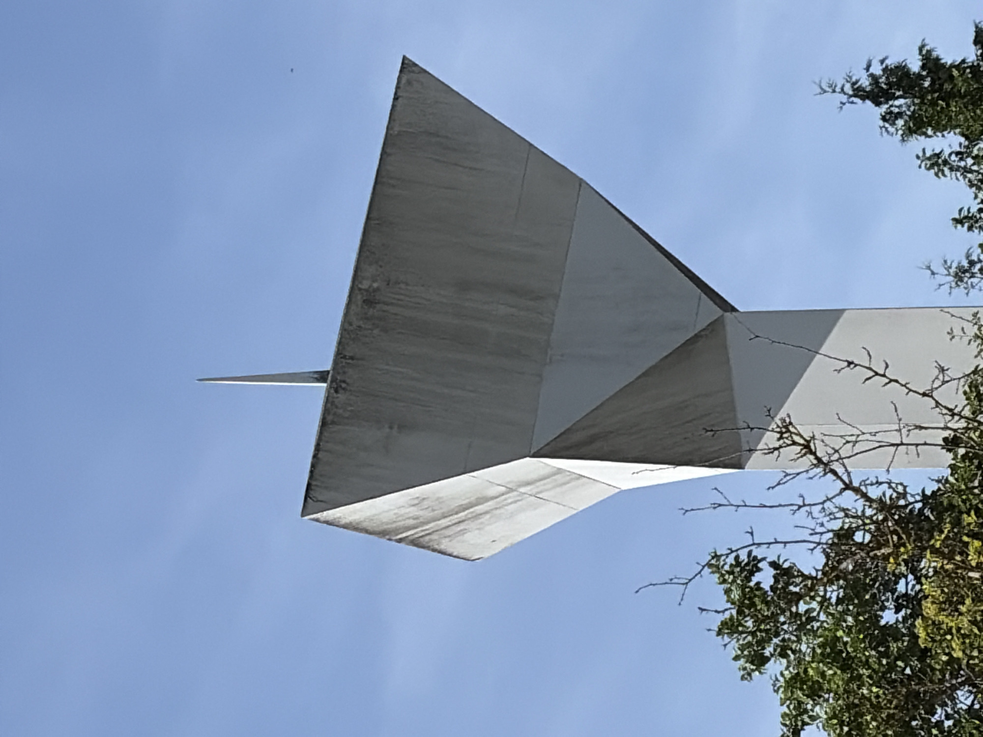 This media file shows a low-angle shot of a modern, geometric sculpture against a clear blue sky. The sculpture is made of light-colored, possibly concrete or metal, and has sharp angles and planes. A thin, pointed element extends from the main body of the sculpture towards the left. The surfaces of the sculpture show some weathering and staining, particularly on the underside.
To the right of the sculpture, branches of a leafy green tree extend into the frame, with some bare twigs visible among the foliage. The lighting suggests it is daytime, with the sun casting shadows that are not clearly visible from this perspective. The overall scene is static, focusing on the architectural form of the sculpture and its interplay with the natural elements of the tree and the sky. No people, text, or specific location cues are visible in the image. The mood is serene and perhaps a little stark, emphasizing the clean lines of the sculpture against the vastness of the sky.