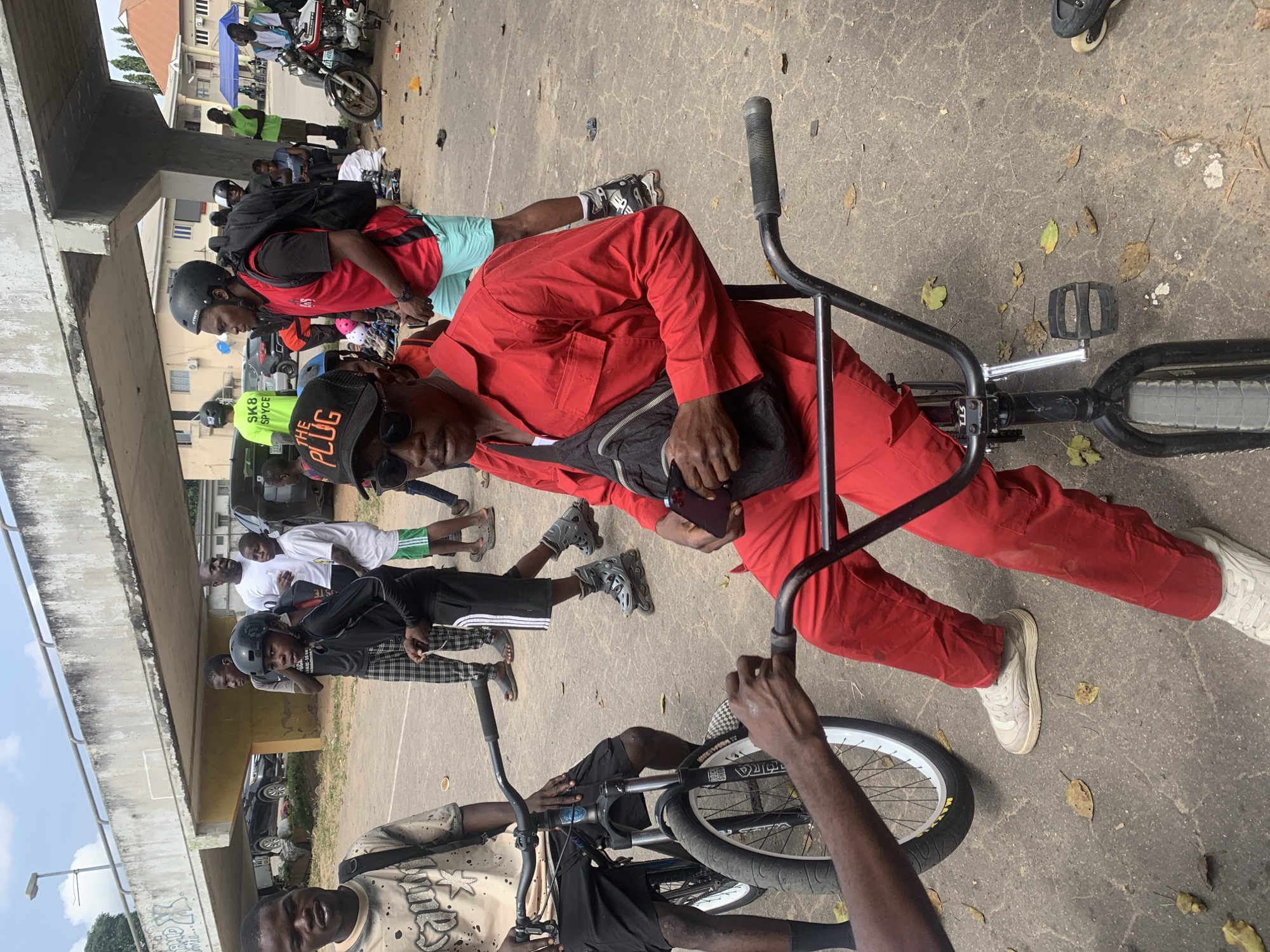 An outdoor scene in Alagbede, Nigeria, features a group of young men gathered under a concrete overpass. The foreground is dominated by a man seated on a BMX bicycle, wearing a red jumpsuit and a black fanny pack. His face is obscured by sunglasses and a black baseball cap that reads "THE PLUG." He holds a smartphone in his right hand. To his left, another young man, wearing a light-colored t-shirt and black shorts, stands with his arm outstretched towards the handlebars of a bicycle.
In the middle ground, several other individuals are visible. One young man wears a black helmet and a red vest over a black shirt, with a large backpack. Another wears a bright green t-shirt with "SK8 SPYCE" printed on it, also sporting a helmet. Several people are wearing rollerblades. A motorcycle is parked in the background to the right, near a blue tent. The ground is paved and appears somewhat dusty, with scattered leaves. Cars and buildings are visible in the background. The overall atmosphere suggests a gathering of youth engaged in outdoor activities, potentially related to cycling and extreme sports.