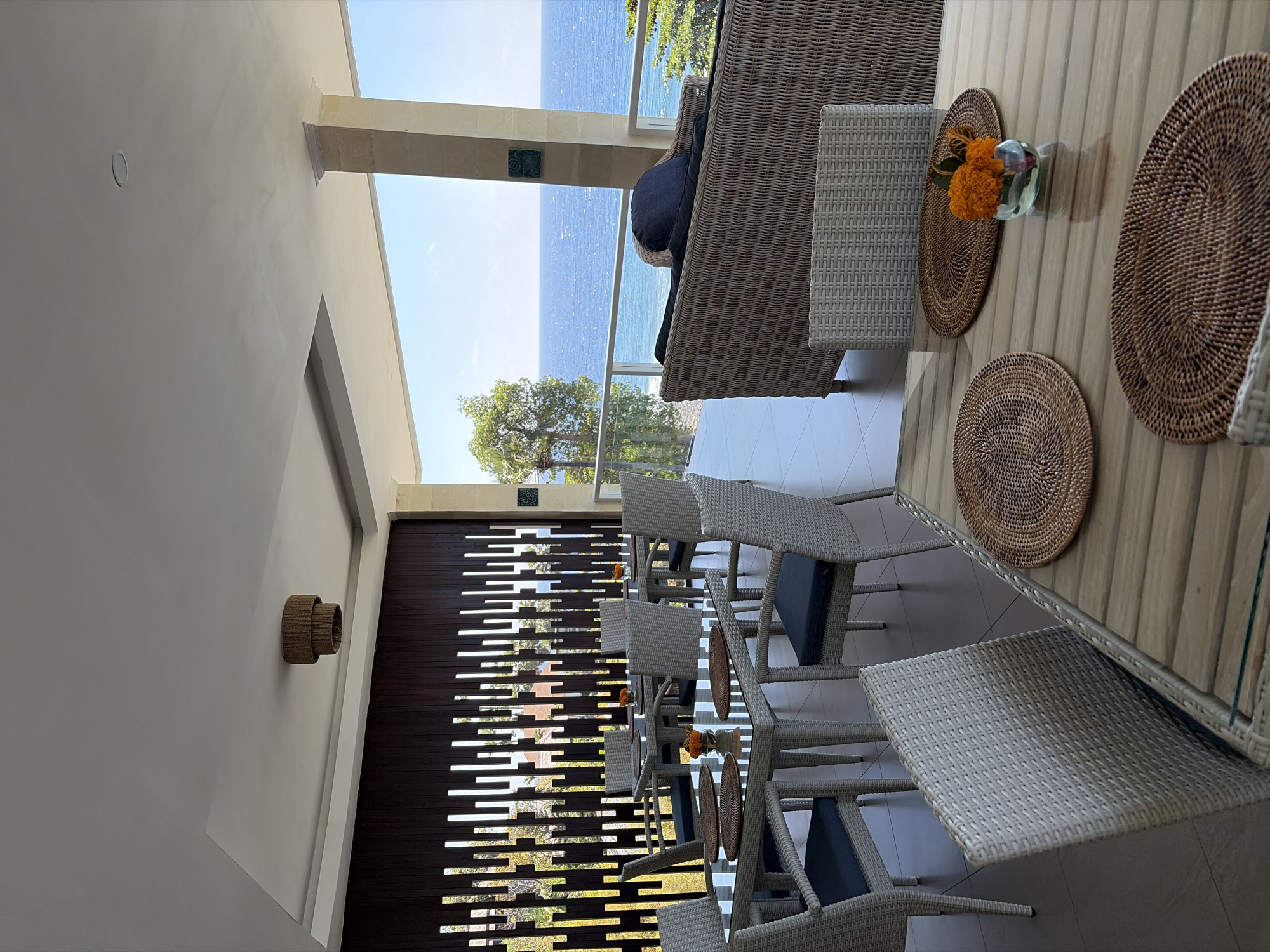 This image depicts an outdoor dining area with a view of the ocean, likely a restaurant or cafe in Abang, Indonesia. The setting is an open-air patio, with a ceiling overhead and a railing overlooking the sea. The foreground features several dining tables, each set with woven placemats and a small vase containing orange flowers. The tables are made of light-colored wood with glass tops, and are surrounded by chairs constructed of woven material with blue cushions.
In the mid-ground, more dining tables and chairs are visible, arranged under a slatted wooden wall. Beyond this, a large woven sofa with dark blue cushions is positioned on the patio. The background is dominated by a panoramic view of the ocean under a clear blue sky. Lush green trees are visible on the coastline to the right. The architectural elements suggest a modern, resort-style establishment. There are no people visible in the image.