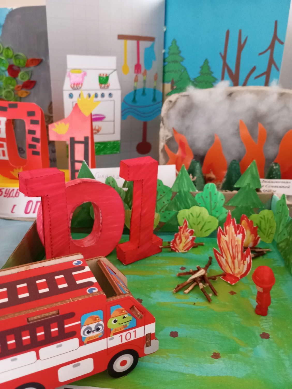 This image captures a vibrant, handmade diorama focused on fire safety education, likely created by children. The display is divided into two primary sections: an outdoor forest scene and an indoor kitchen setting.
The outdoor section, occupying the right and central foreground, features a green base representing grass. Several small piles of twigs are scattered across it, accompanied by paper cutouts depicting bright orange and red flames, indicating various fires or campfires. Paper trees and bushes in shades of green line the back of this section, leading to a backdrop illustrating a blue sky with stylized brown tree trunks. A more significant forest fire is depicted with larger orange flames and fluffy white cotton wool representing smoke, surrounded by grey, rock-like structures. In the immediate foreground, a red toy fire truck with cartoon raccoon and frog characters in the windows, marked with "101," is positioned as if arriving at the scene. A small red figurine of a firefighter stands nearby. Prominently placed in the center foreground are large, red 3D numbers spelling out "101," which is Russia's emergency number for fire services.
The left side of the diorama showcases an indoor kitchen scene. A white cardboard cutout depicts a stove with pots, a sink, and a table adorned with a birthday cake and candles. Hanging kitchen utensils (a spatula, whisk, and oven mitt) are also visible. A small flame cutout near the stove suggests a potential cooking hazard. Further to the left, a partially visible backdrop features a decorative element made with green quilled paper.
The overall impression is a brightly lit, educational display designed to teach about fire hazards in both domestic and natural environments, and the importance of calling the emergency number "101." The craftsmanship indicates it's a school project or a similar creative exhibit.
Visible text includes "101" on the fire truck and the large 3D numbers. Partially visible Cyrillic text in the background includes "Правила безопасн..." (Rules of safet...) likely pertaining to fire safety, and another sign mentions "Будь с огн..." (Be with fire...).