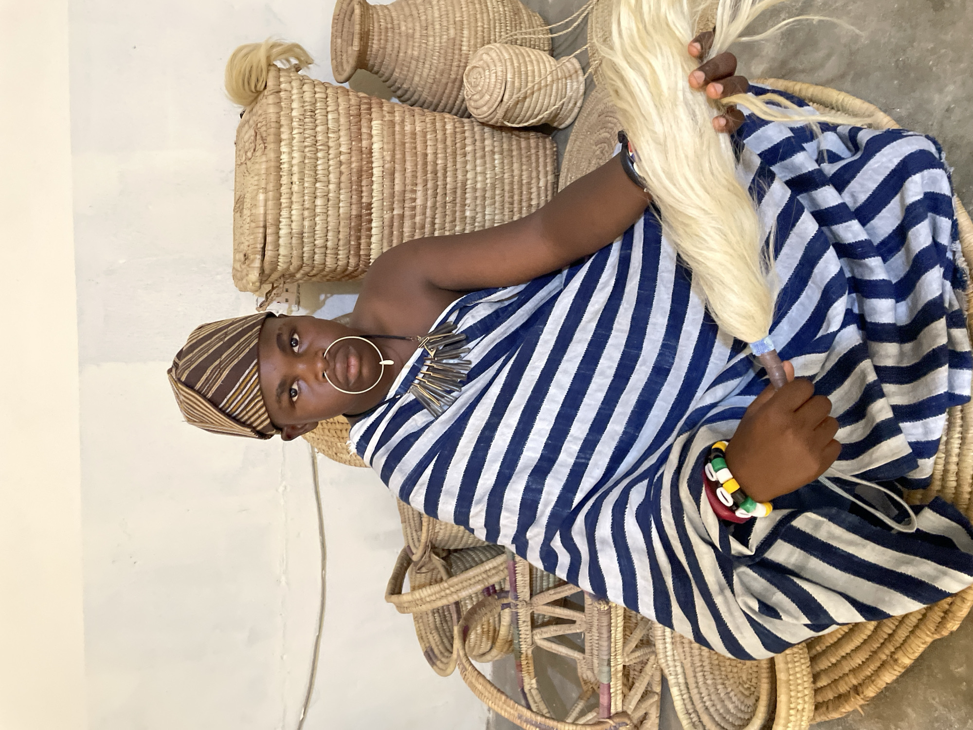 A person with brown skin and dark hair, wearing a striped blue and white cloth, a striped brown cap, a large gold hoop earring, and a necklace of silver pendants, sits in a woven basket chair. The person holds a bundle of light-colored fibers in their hands. Behind them are several woven baskets of various sizes and shapes. The background is a white wall. The location is Jos, Nigeria.