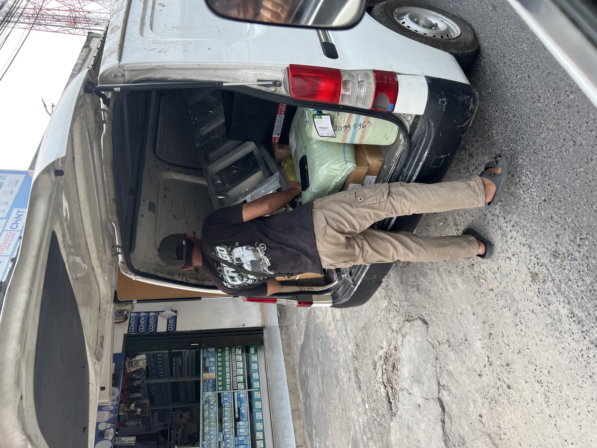 A man wearing a black t-shirt and tan cargo pants stands in the back of a white van, loading packages. The van's rear doors are open, revealing several boxes and bundles inside. To the left of the van, a store with shelves displaying various items is visible. The ground is paved with asphalt. The scene appears to be in a commercial area in Citeureup, Indonesia, as indicated by the presence of the store and the surrounding infrastructure.