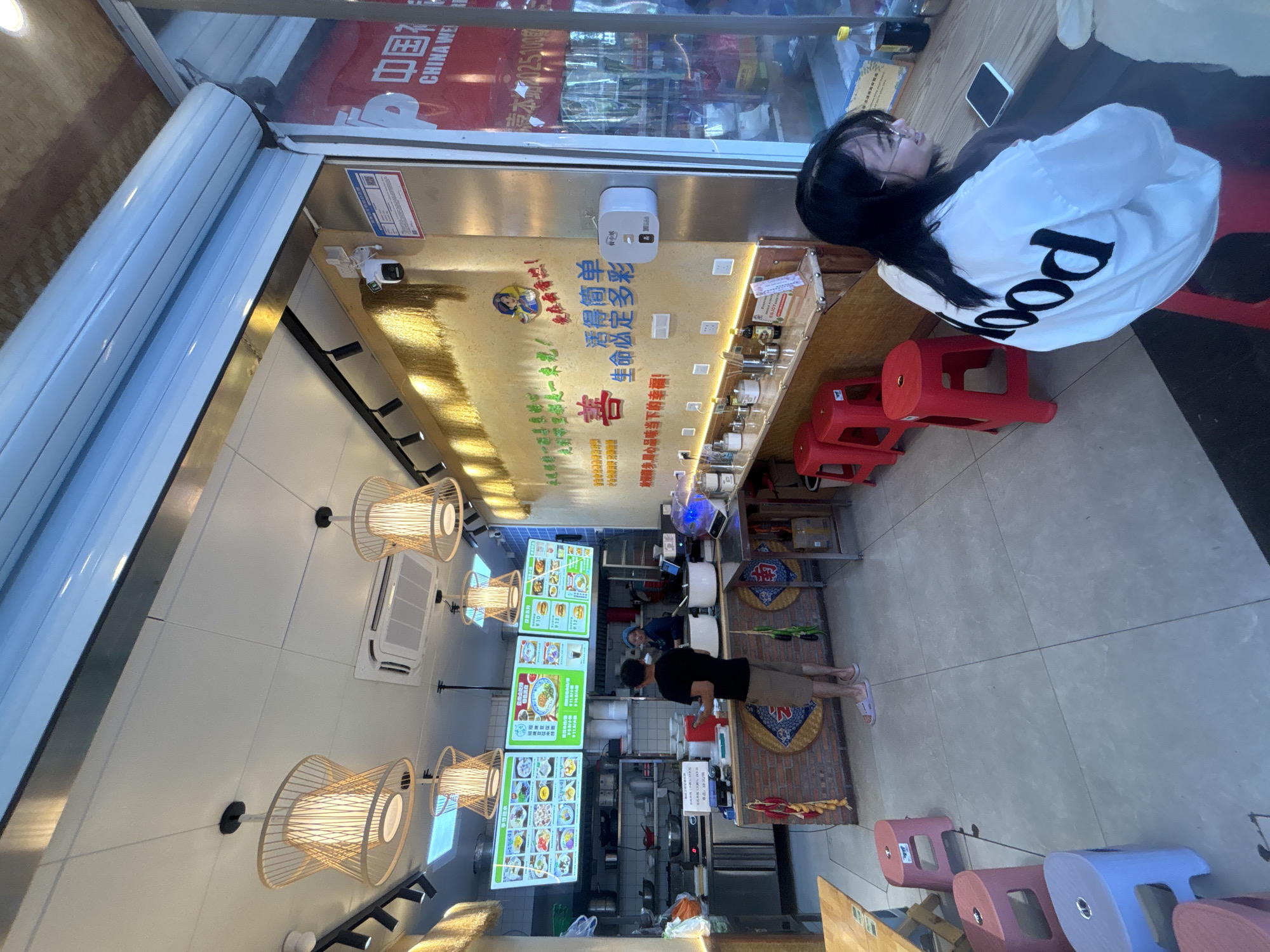 This is a medium shot, taken from a slightly low angle, of the interior of a small restaurant in Suzhou Industrial Park, China. The restaurant appears to be a casual eatery, possibly specializing in noodles or similar dishes, as suggested by the menu boards displayed prominently on the wall.
In the foreground, a young woman with long black hair and glasses is seated on a red stool, facing the right side of the frame. She is wearing a white jacket with the word "food" printed in large black letters on the back. Her right hand is resting on a mobile phone placed on a wooden counter.
Behind her, a man in a black t-shirt and khaki shorts stands at a counter, facing away from the camera, appearing to interact with a food service worker. The worker, wearing a blue hair net, is visible behind the counter, partially obscured by equipment.
The restaurant interior is brightly lit by several pendant lights with bamboo shades hanging from the ceiling. The walls are decorated with colorful menu boards and motivational phrases written in Chinese characters. The counter area is stocked with various containers, bottles, and utensils. Red plastic stools are scattered around the seating area.
The overall environment suggests a clean and functional, yet somewhat cramped, dining space. The image captures a moment of everyday life within a commercial establishment in an urban setting.