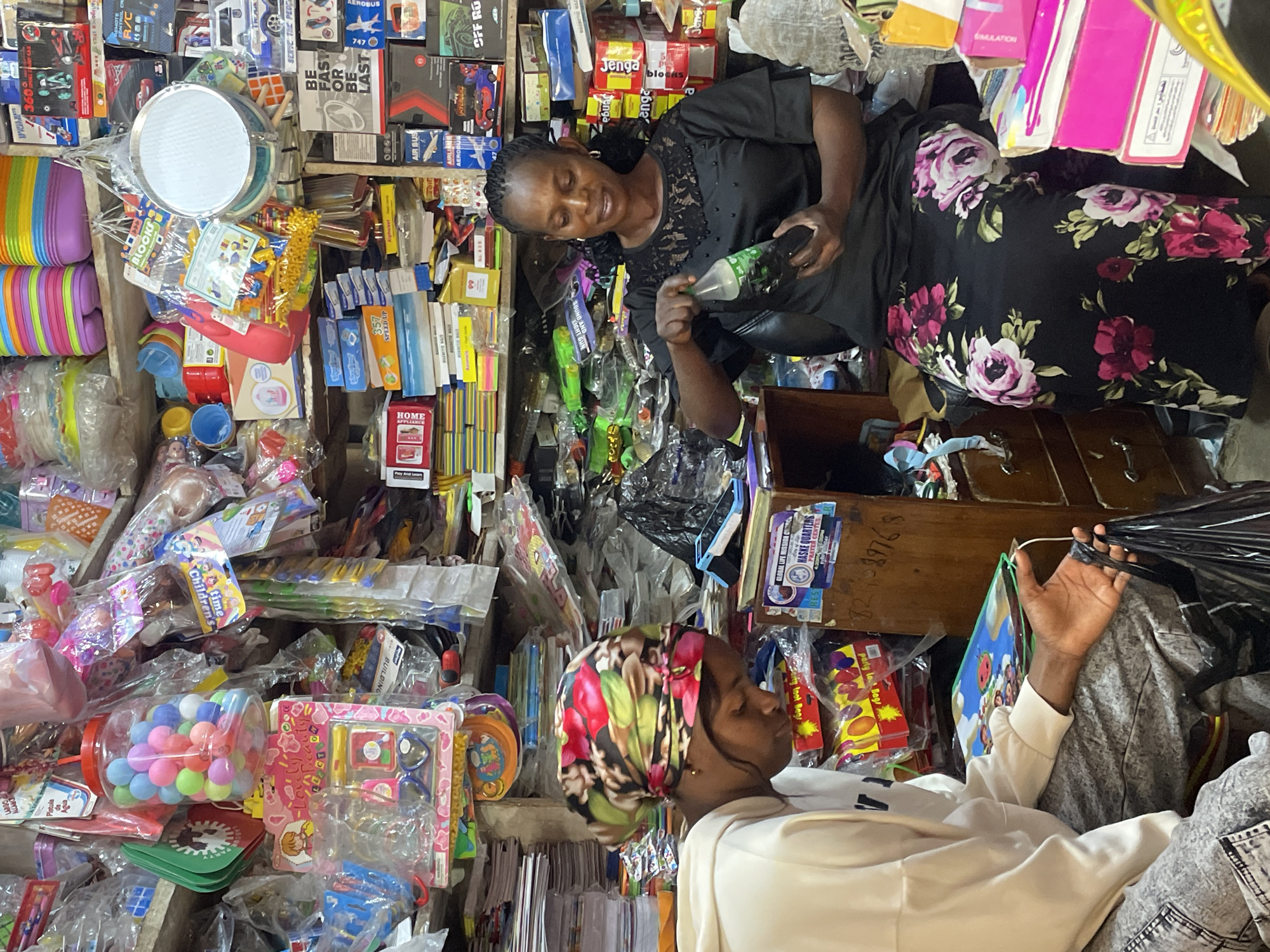 The image captures an indoor scene, likely a shop, filled with various merchandise. Two individuals are present. In the foreground, a young person wearing a white hoodie and grey patterned pants is seated and looking down, holding a black plastic bag. Beside them, a woman in a black dress with a floral pattern and a headscarf is standing, holding a green bottle of soda and appearing to open it. The shelves and surfaces of the shop are packed with a diverse array of items, including toys, stationery, plastic containers, and packaged goods. The overall impression is of a cluttered but functional retail environment. The image is stated to be from Jos, Nigeria.