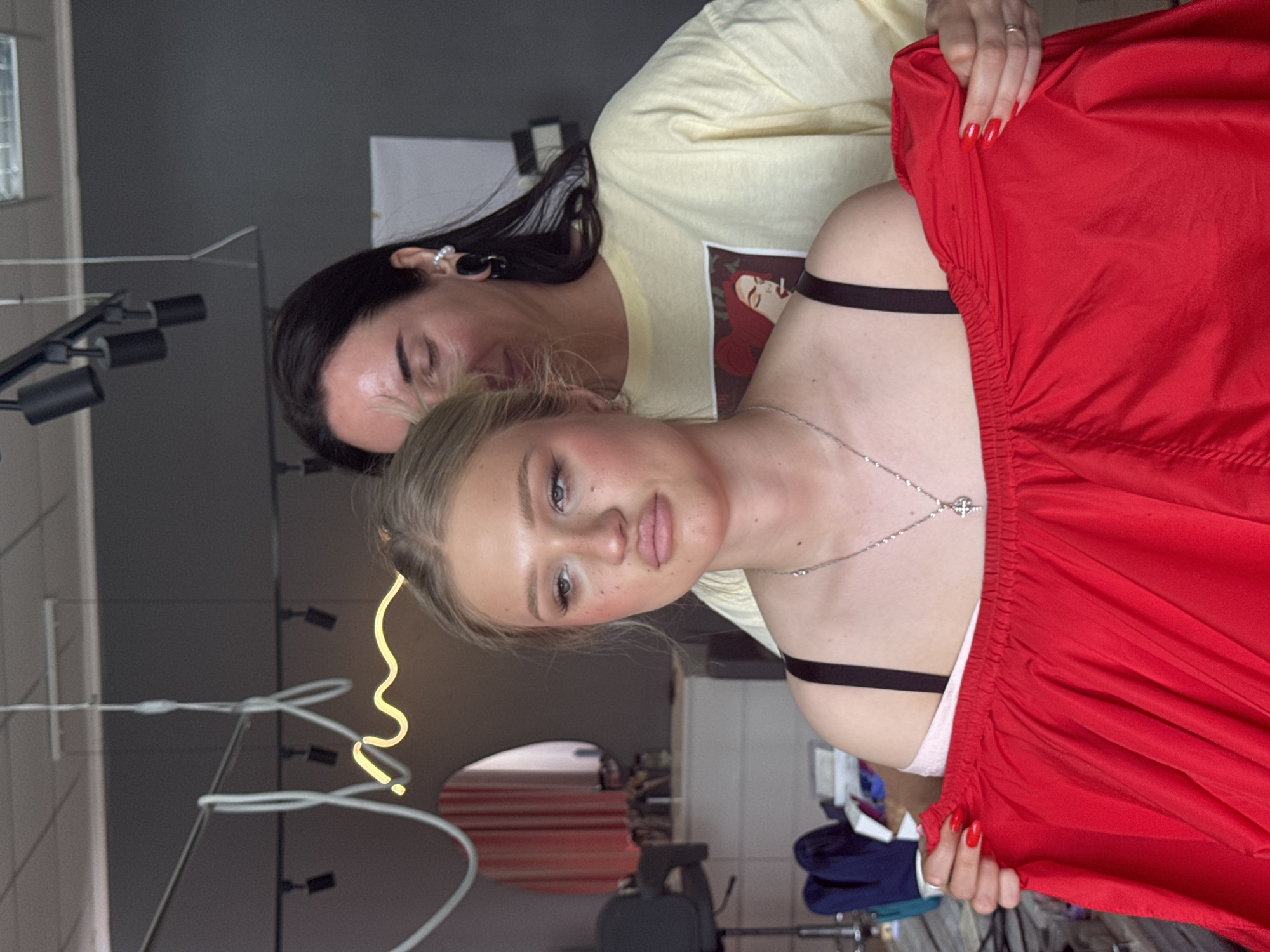 Two women are in a room that appears to be a beauty salon or dressing room in Novocherkassk, Russia. A blonde woman in the foreground, facing the camera with her head slightly tilted, has her lips parted and eyes looking off to the side. She is wearing a black bra with a thin strap and a thin silver necklace with a cross pendant. She is holding a large piece of bright red fabric. Behind her, a dark-haired woman in a pale yellow t-shirt is partially visible, leaning in and looking at the blonde woman. The background shows salon furniture, including a chair and a counter with items on it, as well as a circular mirror with pink curtains. A neon light fixture is also visible hanging from the ceiling.
