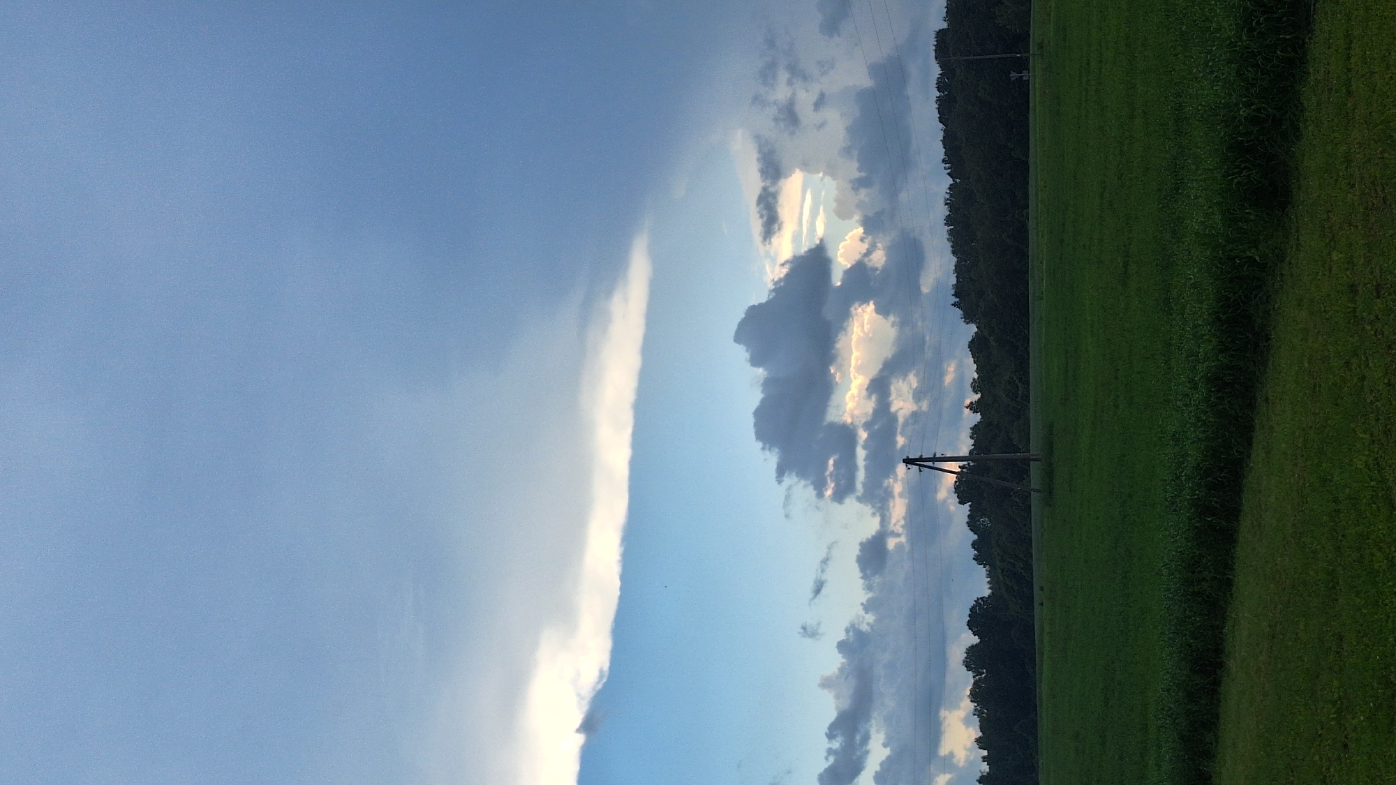 The image shows an overcast sky over a green field. In the middle of the field, there is a wooden pole with wires. A line of trees is visible in the background. The scene appears to be rural, with no buildings or people visible. The sky is a mix of blue and grey clouds, with some sunlight breaking through. The field is lush and green, suggesting it is summer.
