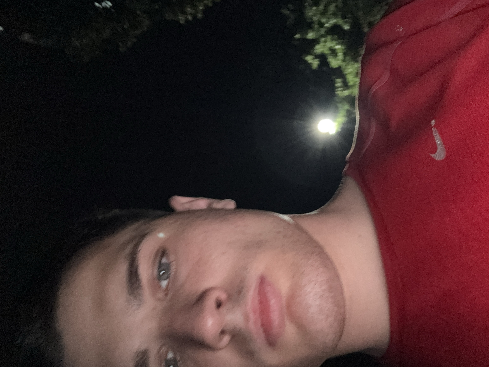 The image is a close-up, low-light selfie of a young Caucasian male. He has fair skin, short brown hair, and is looking directly at the camera with a neutral expression. His eyes are light blue. He is wearing a red t-shirt with a silver Nike logo on the chest. The background is dark, with a bright light source visible in the distance, possibly a street lamp, and some blurred greenery, suggesting an outdoor setting at night. There is no indication of the specific location within Stara Zagora, Bulgaria.