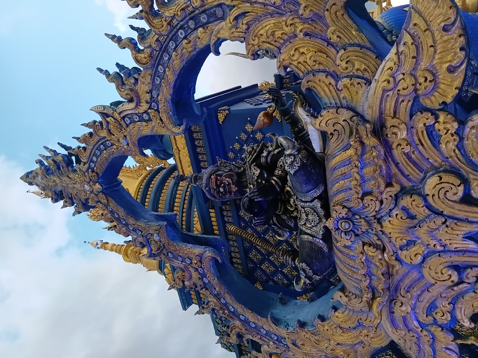 The image shows a low-angle, close-up view of an ornate, blue and gold statue in Chiang Rai, Thailand. The statue depicts a fearsome, dark-skinned mythical creature with an open mouth, sharp teeth, and red eyes. It is seated with one leg bent and the other extended, holding a glass orb in its left hand and a bird figurine in its right. The creature is adorned with intricate, detailed armor and jewelry. Behind the statue, a portion of a Buddhist temple complex is visible, characterized by layered golden spires and blue, gold-accented architecture. The sky above is partly cloudy. The statue is likely a guardian figure at the entrance of the temple, which is part of the Wat Rong Khun (White Temple) or Wat Rong Suea Ten (Blue Temple) complex, both iconic landmarks in Chiang Rai.