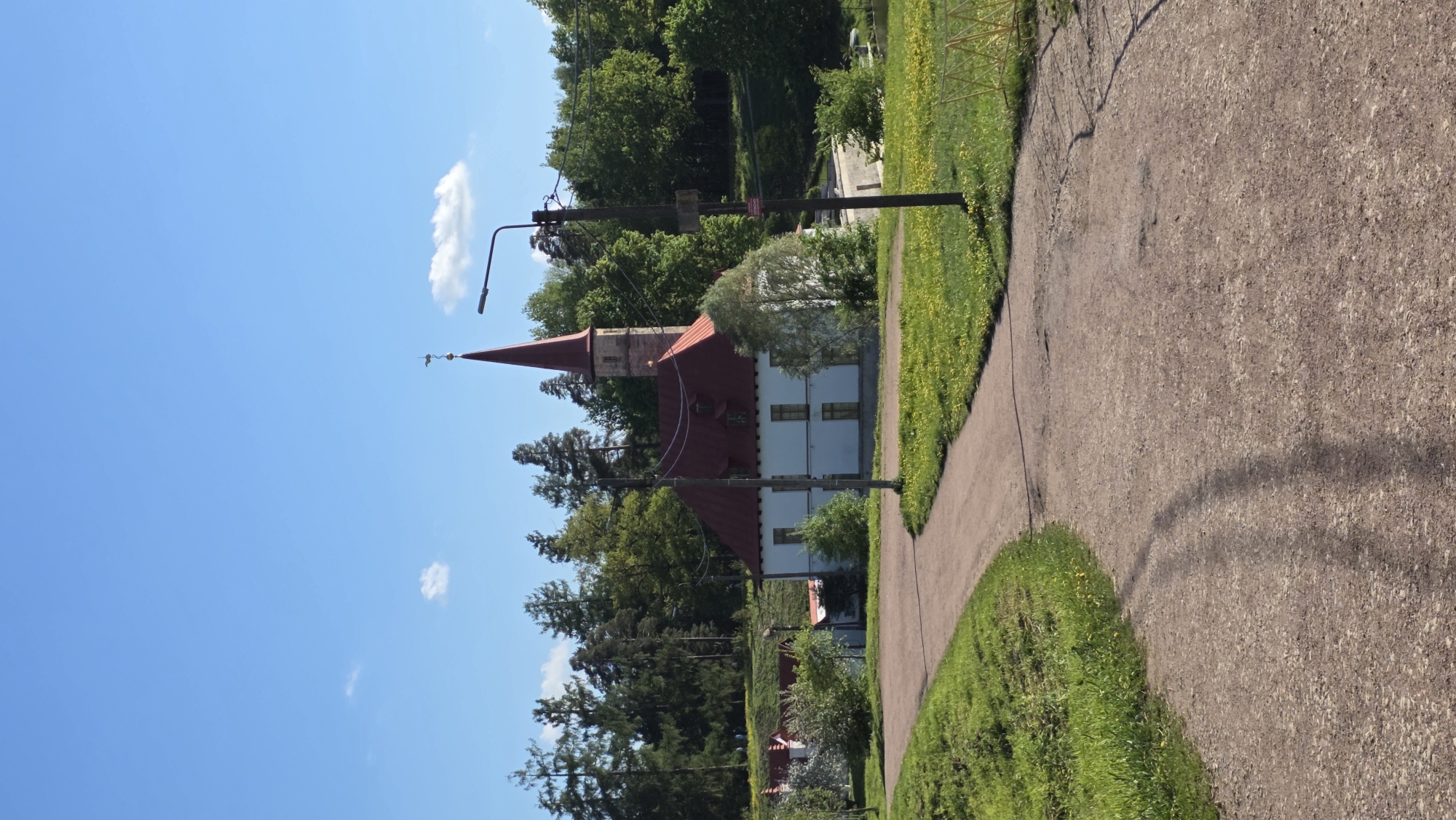 The image depicts a church in Gatchina, Russia, with a red roof and spire, adjacent to a path. The church is framed by trees and a blue sky with scattered clouds. A lamp post stands near the path. The path, with grass on either side, leads away from the viewer.