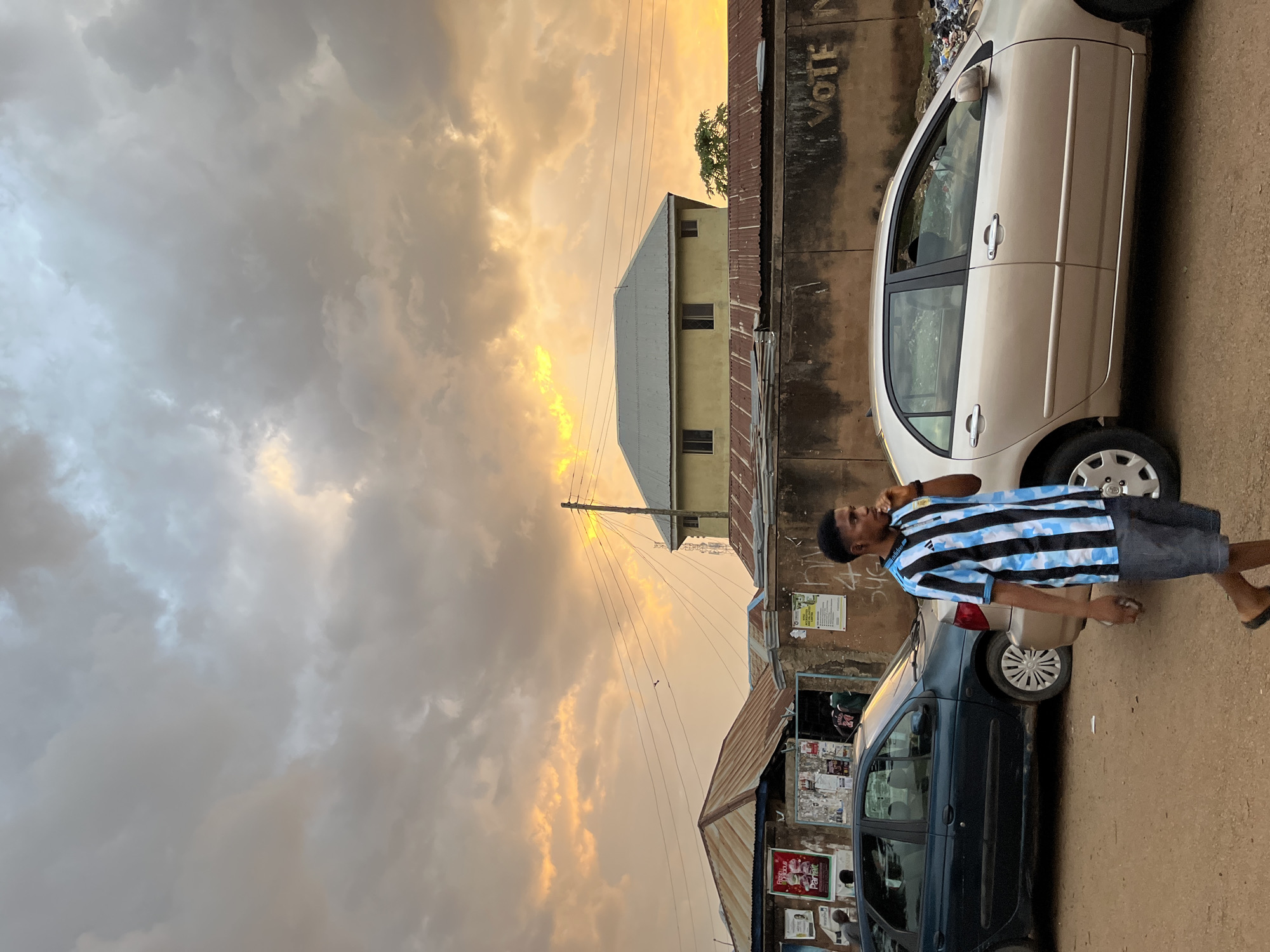 A young Black male walks across a dirt road in Jos, Nigeria, as the sun sets. He wears a blue and white striped t-shirt and gray shorts. Two cars are parked on the right side of the road. A dark green sedan is parked behind him. A lighter colored sedan is parked in front of him. In the background, there are several buildings with corrugated metal roofs. The sky is filled with orange and gray clouds. The scene suggests a residential or commercial area in Jos.