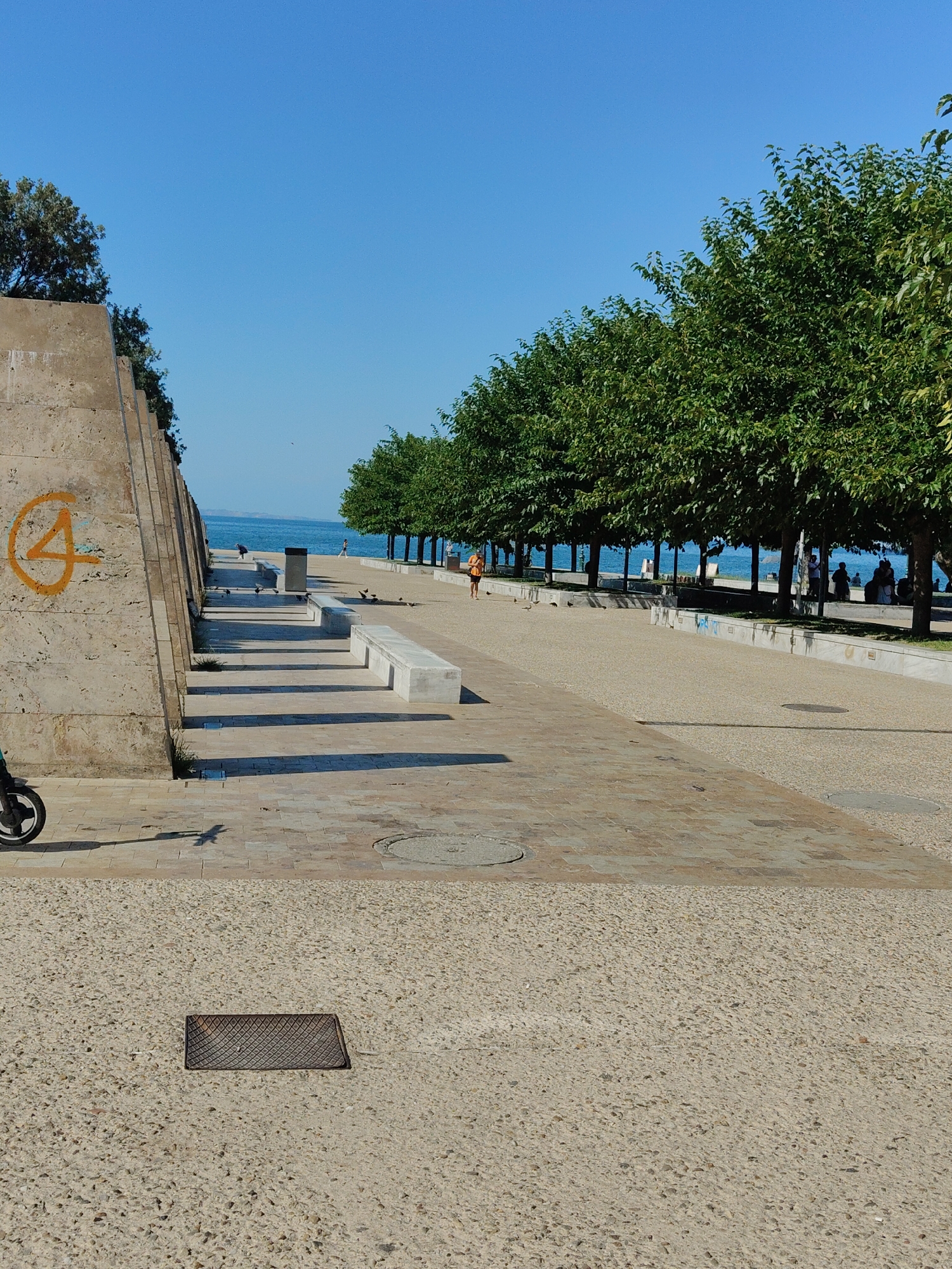 The image depicts a sunlit promenade along the coast of Thessaloniki, Greece. The foreground features a textured concrete walkway with a metal grate. To the left, a segment of a large, angled concrete structure is visible, marked with a yellow spray-painted symbol resembling a spiral with a line. A portion of a black electric scooter is at the bottom left.
The promenade stretches into the distance, lined with a row of deciduous trees casting shadows on the paved surface. Beyond the trees, the blue sea is visible under a clear sky. Several concrete benches are placed along the promenade.
In the middle ground, a person in an orange top is walking away from the viewer. Further along the promenade, a group of people are seated or standing near the trees, closer to the water. A few pigeons are scattered on the walkway. The scene conveys a sense of a relaxed, public space on a bright day.
