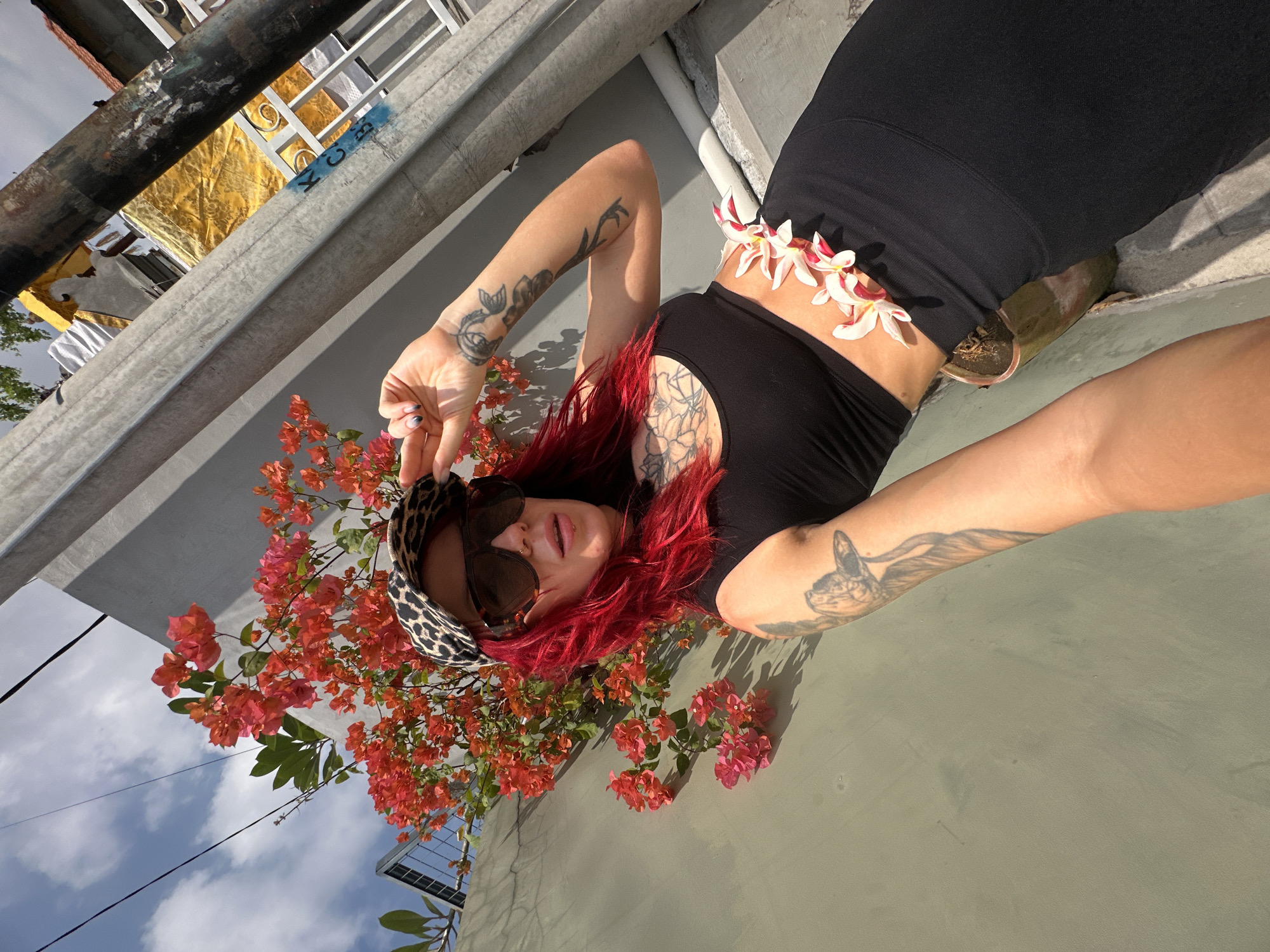 In the city of North Kuta, Indonesia, a woman with bright red hair, wearing a black crop top, black leggings, a leopard print baseball cap, and large sunglasses, takes a selfie outdoors. She is posing against a grey wall with vibrant orange and pink bougainvillea flowers blooming beside her. A lei of white and pink flowers is draped across her waist. Her arms are visible, adorned with tattoos. The background shows a bit of sky with clouds, electrical wires, and what appears to be part of a building with yellow fabric and a white fence. The lighting suggests it is daytime, possibly late afternoon due to the shadows.