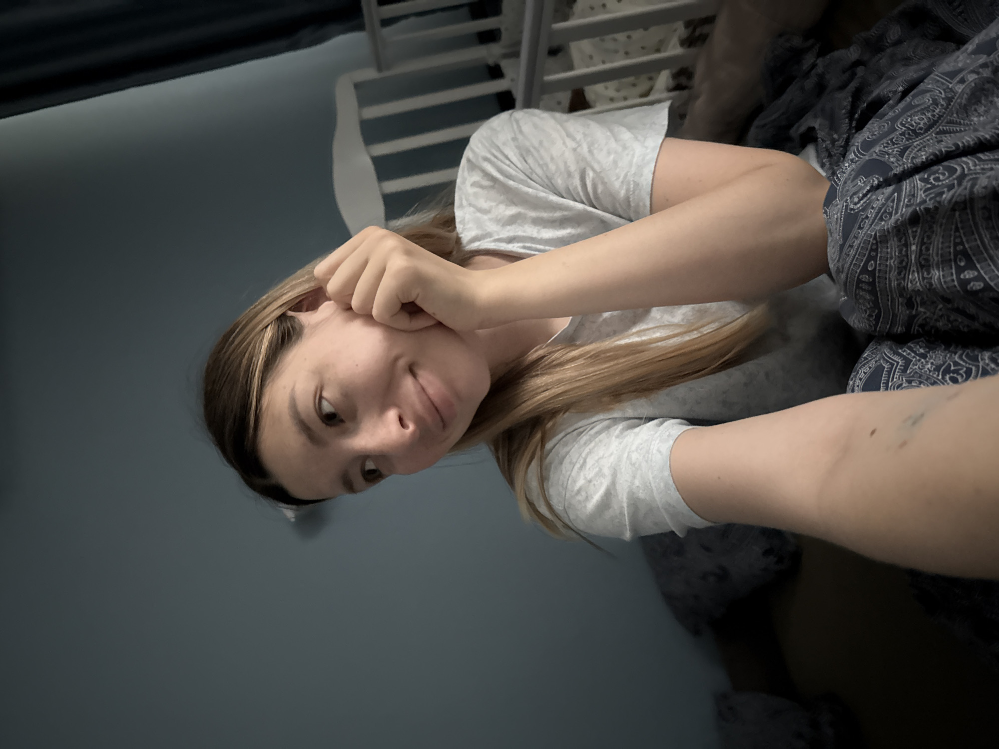 The image shows a young woman with blonde hair sitting on a bed, looking to her left. She is wearing a grey t-shirt and is resting her chin on her hand. A crib and a dark blue curtain are visible in the background. The room is dimly lit, with the blue wall providing a muted backdrop. There is no indication of the location being Moscow, Russia.