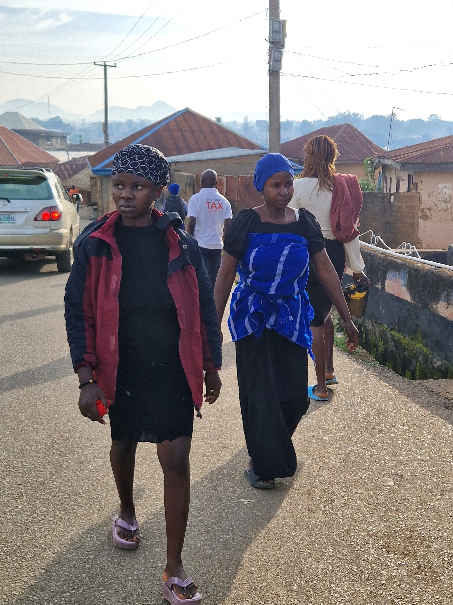 In Kabong, Nigeria, two women walk side-by-side on a paved road in front of buildings with corrugated metal roofs. The woman in the foreground wears a black dress, a maroon and navy jacket, and a patterned headscarf, with a red object in her right hand. She is wearing platform flip-flops. The woman to her right wears a blue headscarf, a black top with a blue and white patterned wrap around her waist, and black pants. She is also wearing flip-flops. Behind them, a man in a white t-shirt with "PAY YOUR TAX" printed on it walks away from the camera. In the background, a silver SUV is parked, and power lines run across the sky. The scene appears to be captured in daylight with hazy sunlight.