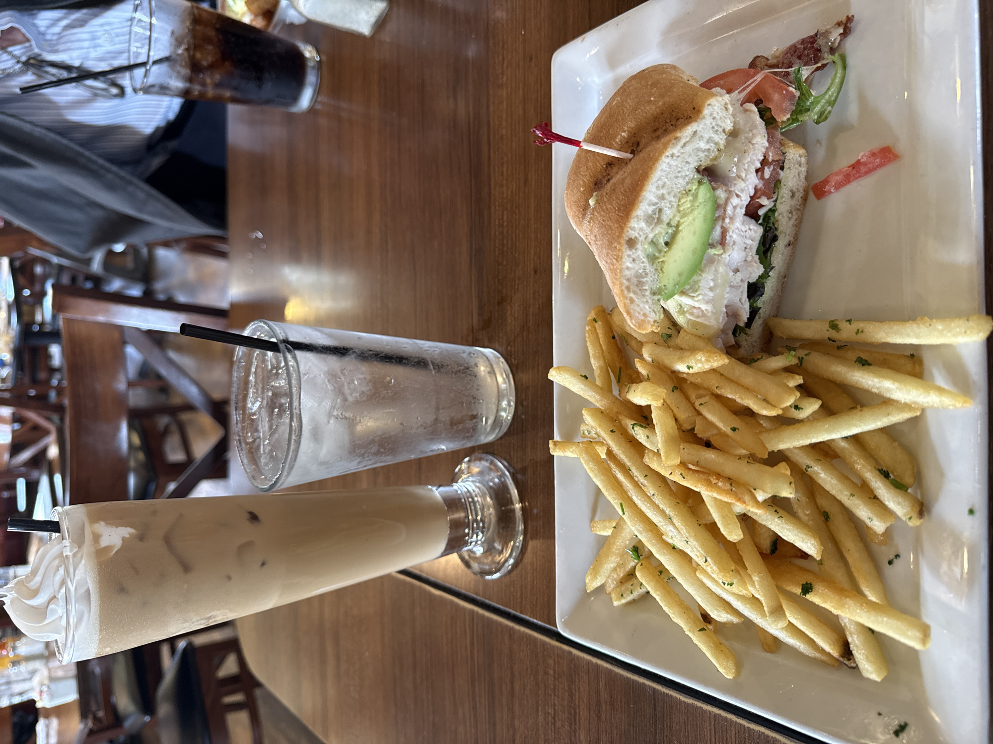 The image shows a meal on a wooden table. The meal consists of a sandwich with avocado, lettuce, tomato, and bacon, accompanied by a portion of French fries. A tall glass of iced coffee with whipped cream, a glass of water with a straw, and a glass of soda can also be seen. The location may be in San Diego, United States of America. No people are fully visible in the image, although the sleeve of a person wearing a blue and white striped shirt is partially visible in the background.