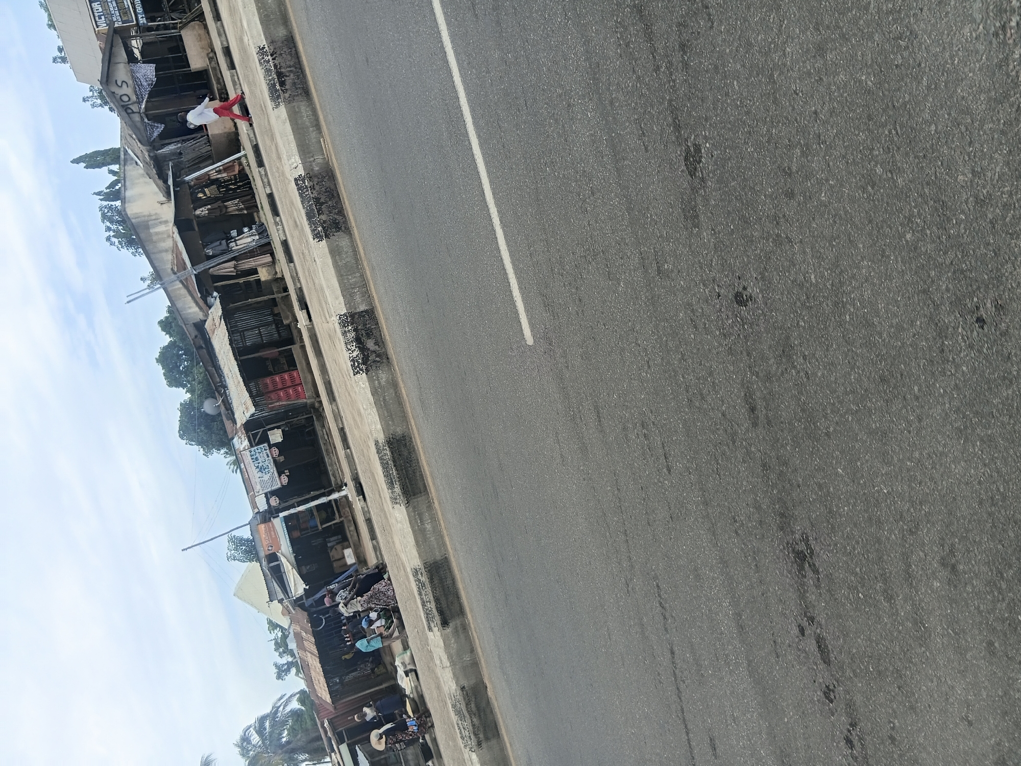 The image depicts a street scene in Jalingo, Nigeria, with a focus on a row of market stalls bordering a paved road. The perspective is angled, with the road occupying the right side of the frame and the market stalls and a stretch of sky to the left. The sky is light blue with some white clouds, suggesting it is daytime, likely morning or afternoon.
In the market area, several stalls with corrugated metal roofs are visible. Some stalls are open, displaying various goods, while others appear closed or under construction. There are multiple people present, though they are not sharply in focus. One individual, wearing a white top and red trousers, is standing near one of the stalls. Other people are seen sitting or standing amongst the stalls, and some appear to be involved in commerce or daily activities associated with a market. Objects visible include crates, textiles, and umbrellas, hinting at the variety of goods sold.
The road is asphalt with a white dividing line. The curb separating the road from the market area shows signs of wear and tear. The overall impression is of a busy, informal marketplace adjacent to a main thoroughfare. The scene suggests a typical day in a Nigerian town, characterized by local commerce and community life. There is no strong indication of any specific event, but rather a snapshot of everyday activity. Text is visible on some of the stall signs, but it is mostly illegible at this resolution, though one sign appears to read "VICTOR" at the top.