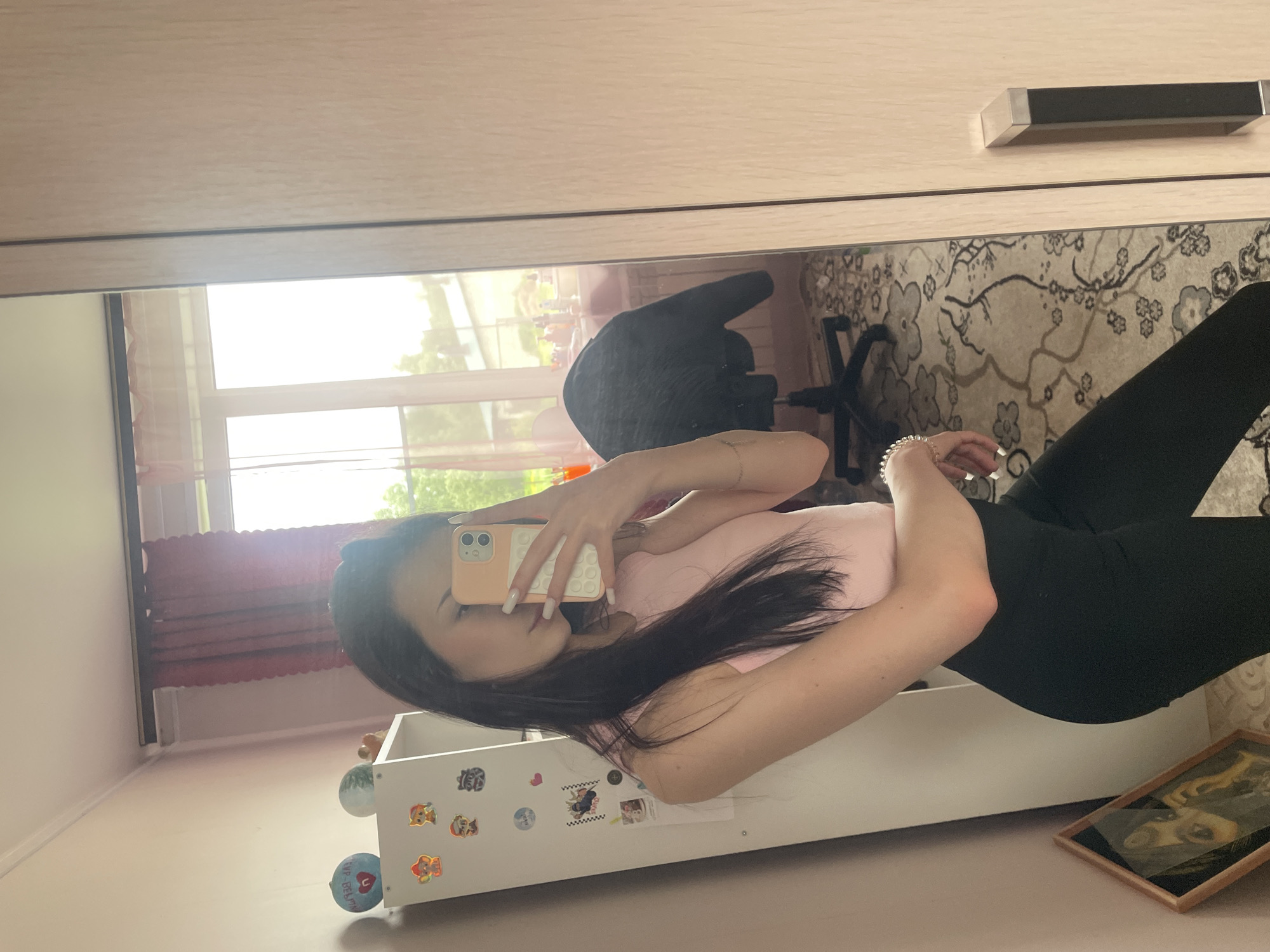 An indoor mirror selfie is captured, showing a person holding a phone, with the image rotated 90 degrees counter-clockwise. The person, likely female, is wearing a light pink top and black leggings. Their face is mostly obscured by the phone, which has a light peach-colored, textured case. Long dark hair falls over their shoulder. White, long fingernails are visible, and the person wears a beaded bracelet on their right wrist.
The setting appears to be a room in a home in Медведево, Russia. In the reflection, a bright window is visible on the left, letting in abundant natural light, suggesting it is daytime and sunny. Reddish sheer curtains are partly visible near the window frame. An office chair with a dark back and wheels is reflected behind the person. On the right, a patterned rug with floral motifs is seen on the floor. A light-colored wooden wardrobe or cabinet with a black and silver handle is reflected at the top of the image.
Below the person, a white rectangular storage unit or shelf is visible, adorned with various stickers, including several Paw Patrol characters, a heart, a checkered flag, and a circular sticker featuring the text "1up-BETA." A small framed picture, possibly an illustration or photo, lies on the floor or a low surface near the bottom right corner of the reflected scene. The floor is light-colored. The overall impression is a casual, personal space.