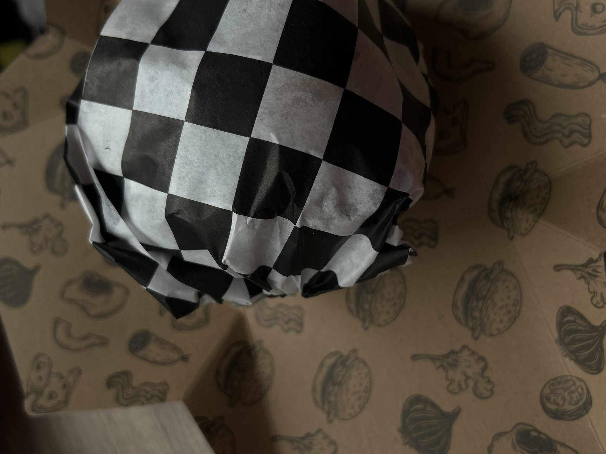 A high-angle, close-up shot depicts a burger wrapped in black and white checkered paper. The wrapped burger is located inside a brown cardboard box, decorated with grayscale drawings of food ingredients, including what appears to be onions, lettuce, bacon, sausages, burgers, eggs, and tomatoes. The box also has a part of a white surface showing in the bottom left. There are no identifiable people, actions, or interactions visible in the image. The location is unknown.