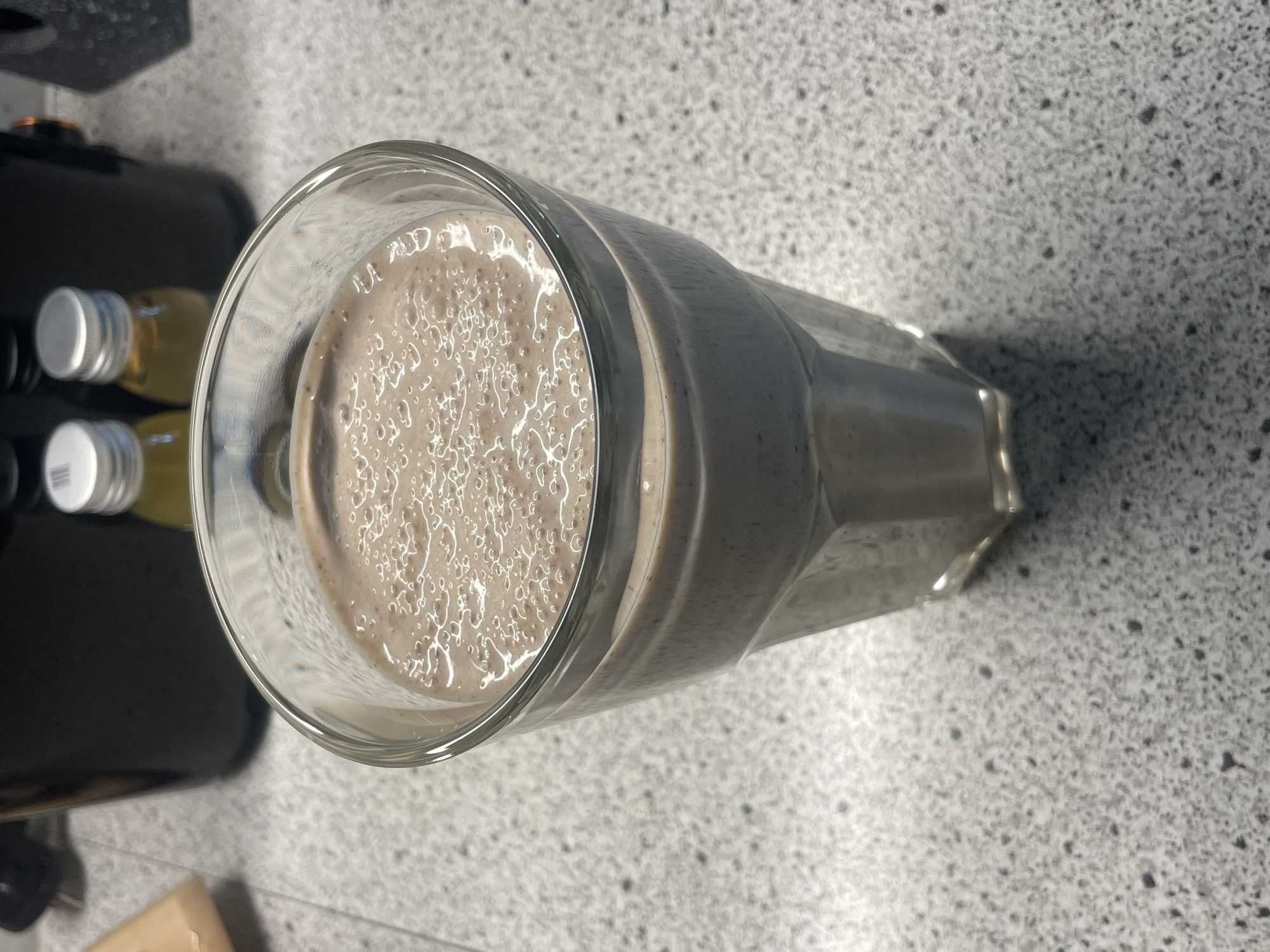 The image shows a glass of chocolate smoothie on a countertop. The glass is transparent and cylindrical, filled with a frothy, light-brown liquid. A few small, clear bubbles are visible on the surface. The countertop has a gray, speckled pattern. In the background to the left, there is part of a black machine and two small bottles, one filled with a yellow liquid, both with silver caps. The location is unknown.