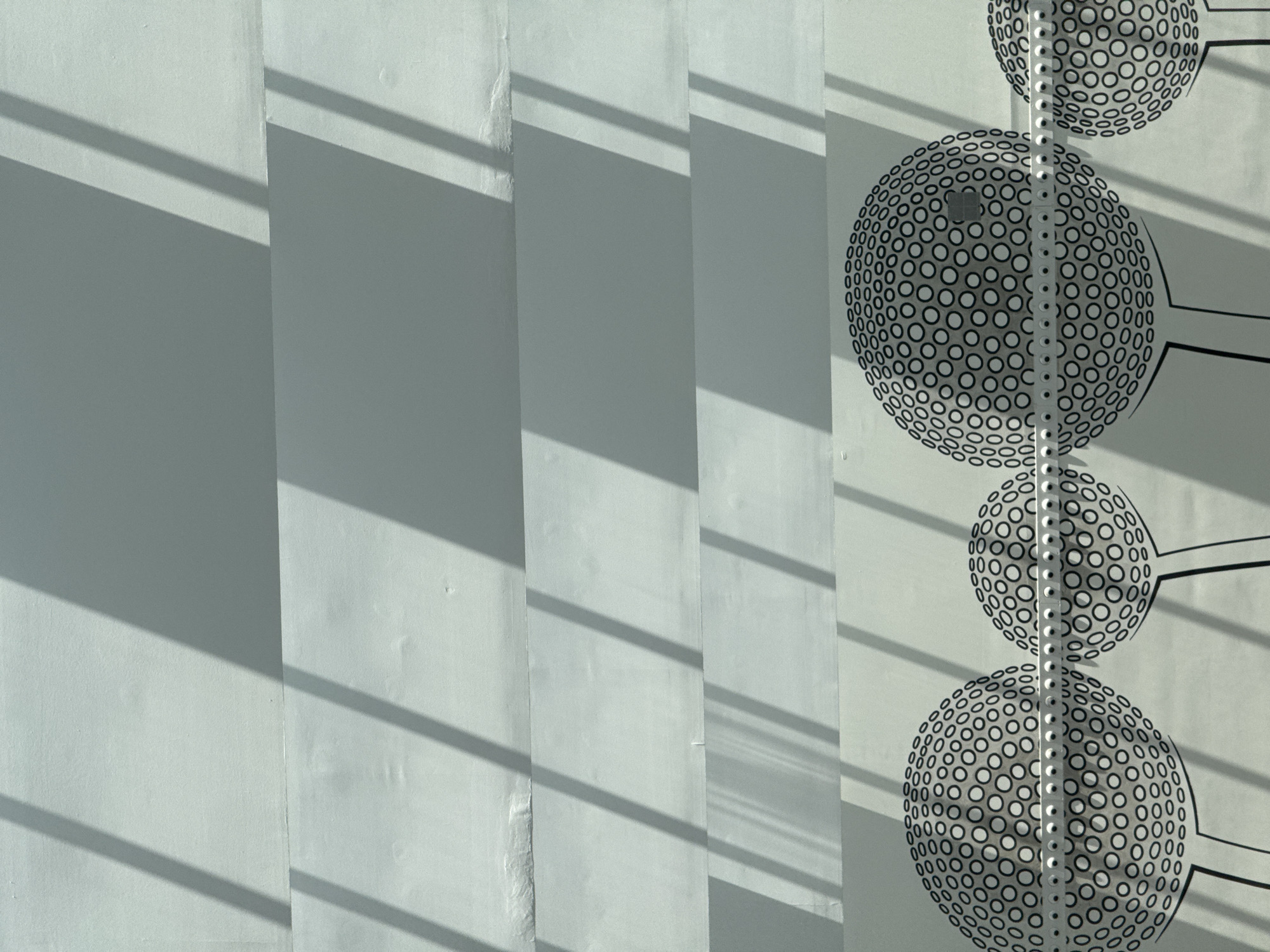 The image is a close-up, high-angle shot of a white wall with diagonal shadows cast across it. The shadows create a geometric pattern on the textured surface. In the lower portion of the image, there is a repeating pattern of black and white circular designs resembling stylized dandelions or golf balls on stems. These designs are applied to the wall, and a horizontal metal fixture with small circular elements runs across them. The wall appears to be outdoors, as suggested by the natural light and shadows. The setting is likely a modern building or outdoor structure. It is not possible to definitively determine the exact location within Majadahonda, Spain based solely on this image.