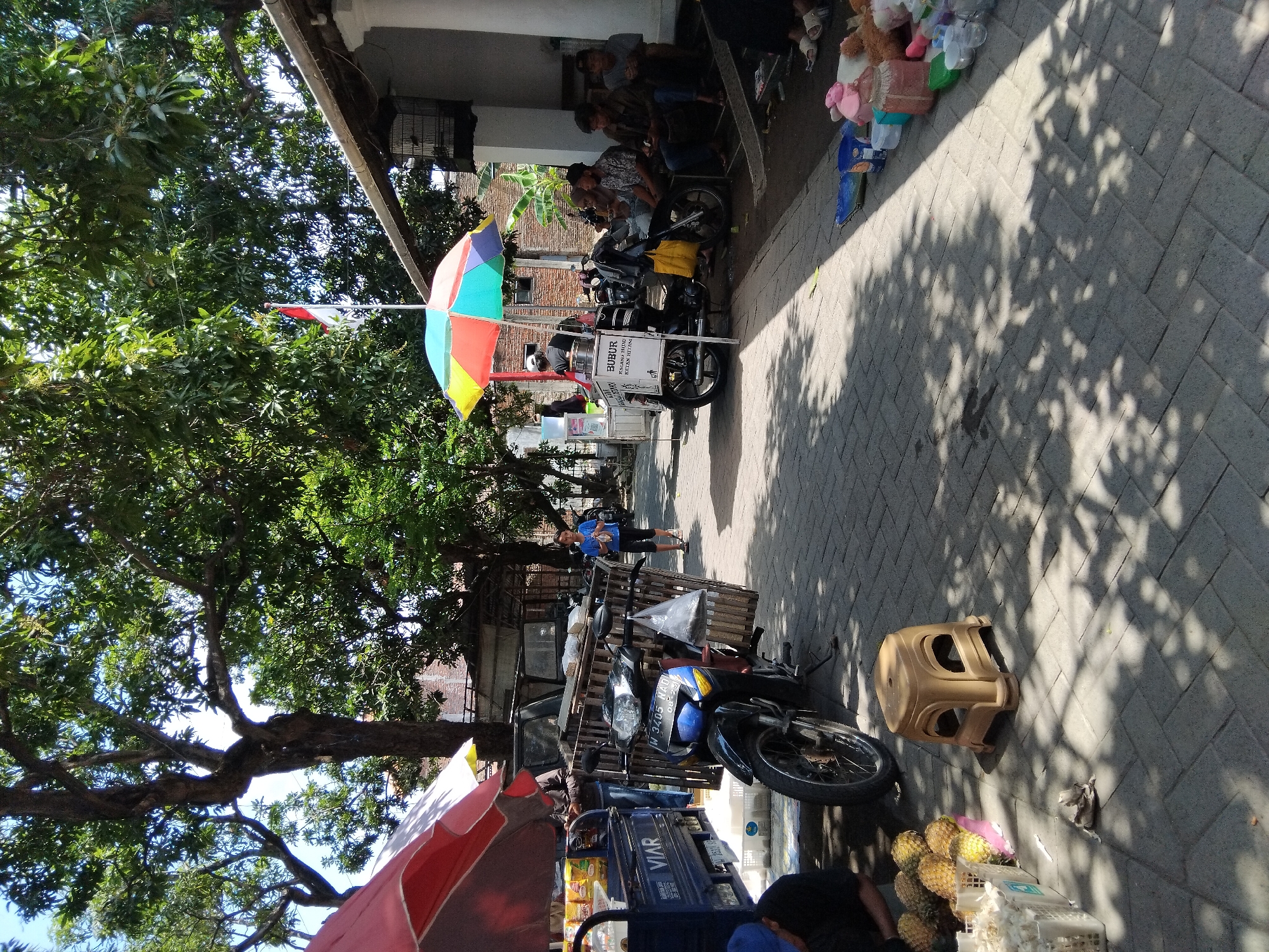 This is a medium-shot, eye-level photograph capturing a street scene in Kepuh Kiriman, Indonesia. The perspective is from a slightly elevated position, looking down a paved walkway lined with various stalls and people.
In the foreground, to the left, a person wearing a blue cap is partially visible, seated next to a cart filled with pineapples and other produce. A tan plastic stool sits on the paving stones. To the left of this, a blue cart with the "VIAR" logo is parked, containing stacked items. A motorcycle with a wooden cage attached to the back is parked in the center of the frame, its license plate reading "W 3405 HAO."
Further down the walkway, a brightly colored, multi-panel umbrella shades a food stall. A person is visible behind the counter of this stall, and another individual, a young woman in a blue t-shirt, walks away from it. Several men are seated on a low platform to the right of the food stall, appearing to be customers or loiterers.
In the background, more stalls and buildings are visible, along with lush green trees providing shade. A red and white flag is flying from a pole above the food stall. The paving stones are a mix of gray and lighter tones, with shadows cast by the trees and structures creating a dappled pattern across the scene. The overall atmosphere appears to be that of a busy market or roadside vendor area on a sunny day.