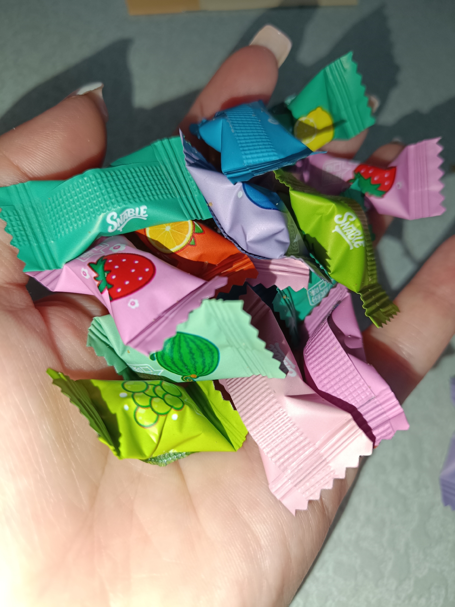 A hand holds a pile of "SNAQABLE" brand wrapped candies. The candies are various colors including light pink, dark pink, green, light green, orange, blue, and purple. Each wrapper has a fruit image on it. The scene is indoors, in natural light. The location is likely not Tobolsk, Russia.
