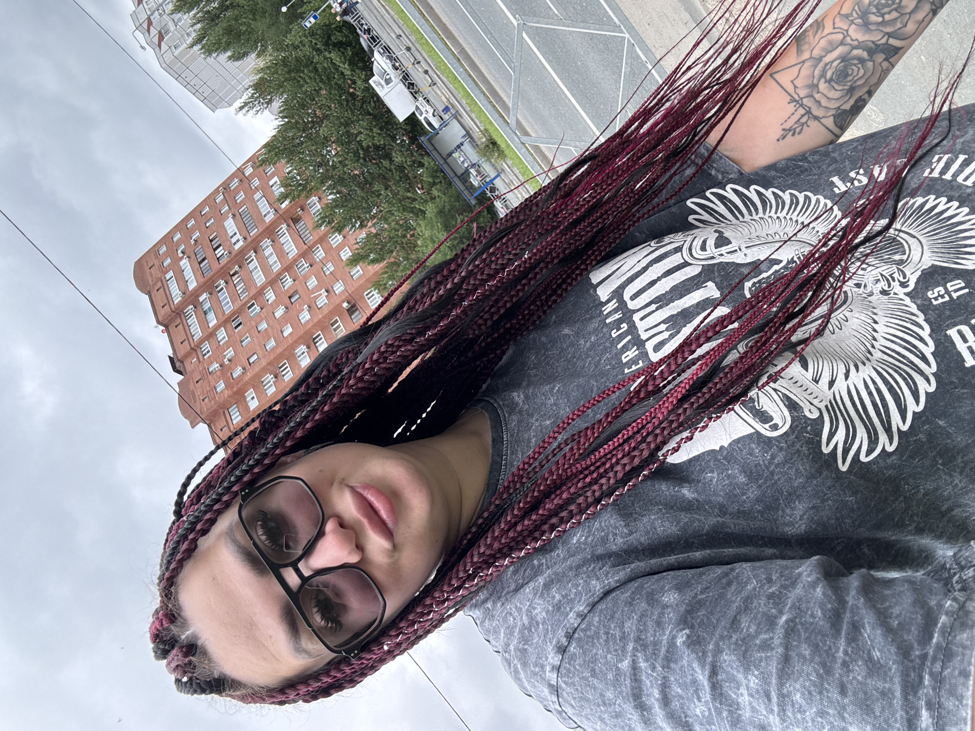 A woman with dark braided hair, interwoven with burgundy strands, takes a selfie outdoors. She is wearing large black-framed sunglasses and a grey t-shirt with a graphic print. Her left arm, visible in the frame, features a tattoo of a rose and geometric shapes. In the background, a tall red brick apartment building, characteristic of Tolyatti, Russia, stands against a cloudy sky. Trees and a street with a road, sidewalk, and parked vehicles are also visible.
