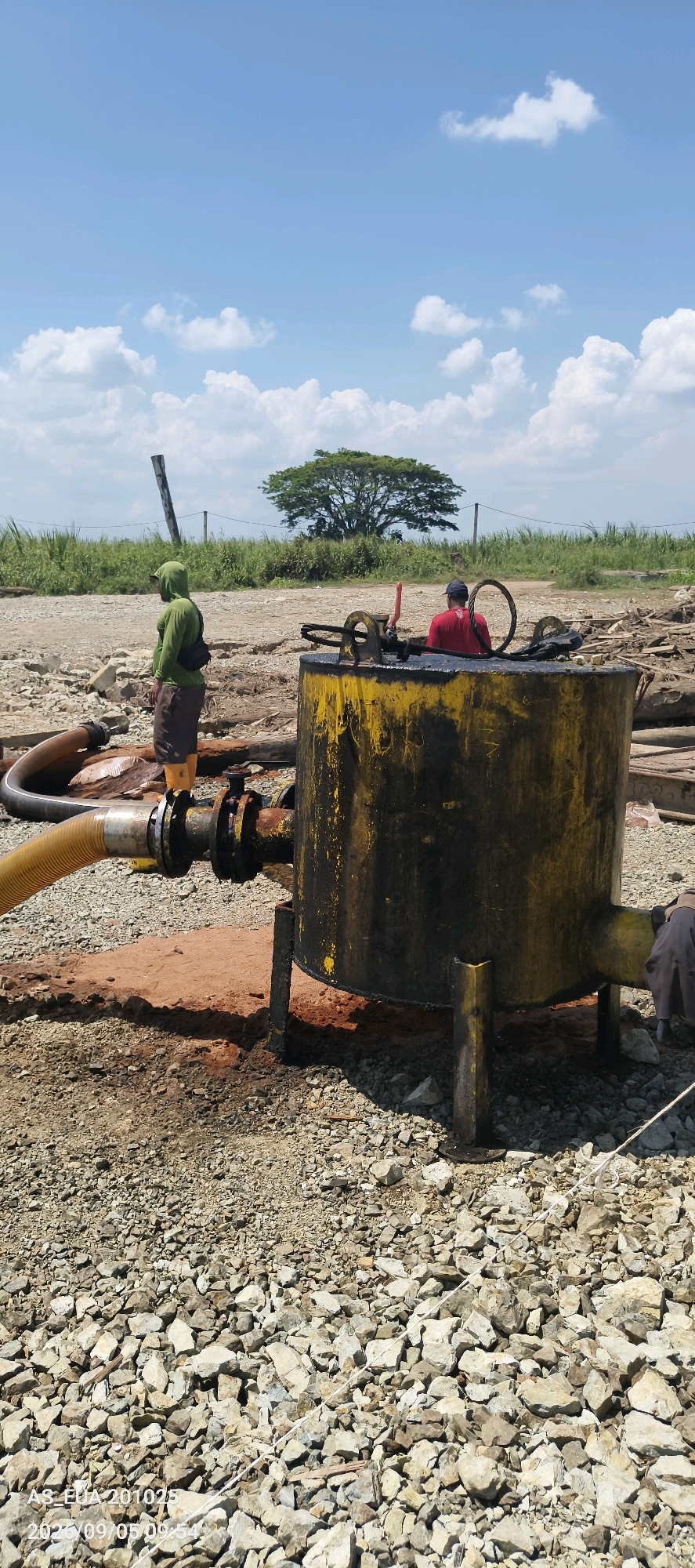 The image depicts an outdoor scene with two individuals working near a large, yellow, cylindrical metal tank. The tank is positioned on a rocky, gravelly surface, and appears to be part of an industrial or construction operation. A thick yellow hose is connected to the side of the tank, leading away to the left. The ground surrounding the tank is a mix of brown earth and grey rocks.
One person, wearing a green hooded jacket and dark shorts with yellow boots, stands to the left of the tank, looking away from the camera. They have a black bag slung across their chest. The second person, wearing a red shirt and a dark baseball cap, is visible in the background behind the tank, appearing to interact with the machinery on top. A large tree with a full canopy stands in the mid-ground, behind a line of green vegetation and a wire fence. The sky above is blue with scattered white clouds. The timestamp "2023/09/05 09:54" is visible in the lower left corner of the image, along with "AS_ELIA201025".
The scene is likely a remote or undeveloped area, possibly a construction site or a processing facility, given the heavy machinery and the natural, unmanicured surroundings. The specific location in Kanduanum 2, Papua New Guinea, suggests this might be related to mining, resource extraction, or agricultural processing, common activities in that region.