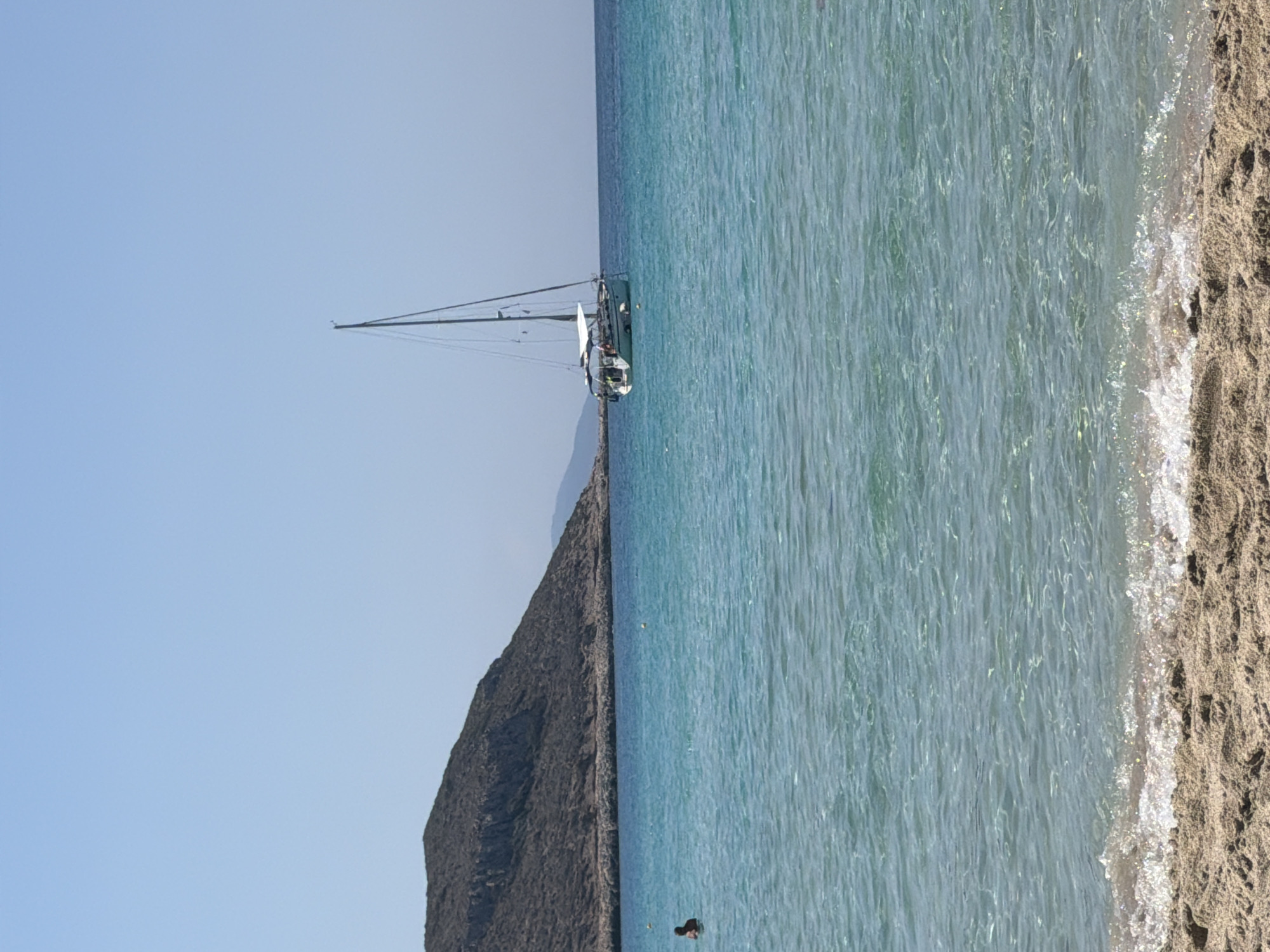 This is a medium shot, eye-level photograph taken on a clear, sunny day at an unknown beach location in Greece. In the foreground, the sandy beach slopes down to the crystal-clear, turquoise water. Gentle waves are breaking on the shore, with sunlight glinting off the water's surface.
In the middle ground, a person is visible in the water on the left side of the frame, appearing to be swimming or wading. Further to the right, a white sailboat is anchored. The boat has two masts and several sails are furled. A white canopy covers part of the deck.
In the background, a large, rocky, treeless hill dominates the left side of the frame. The hill is covered in dry vegetation and appears to be part of the coastline. The horizon line is clearly defined between the sea and the pale blue sky. The water is calm and transparent, allowing a view of the sandy seabed in the shallower areas. The overall impression is one of a serene and picturesque coastal scene.