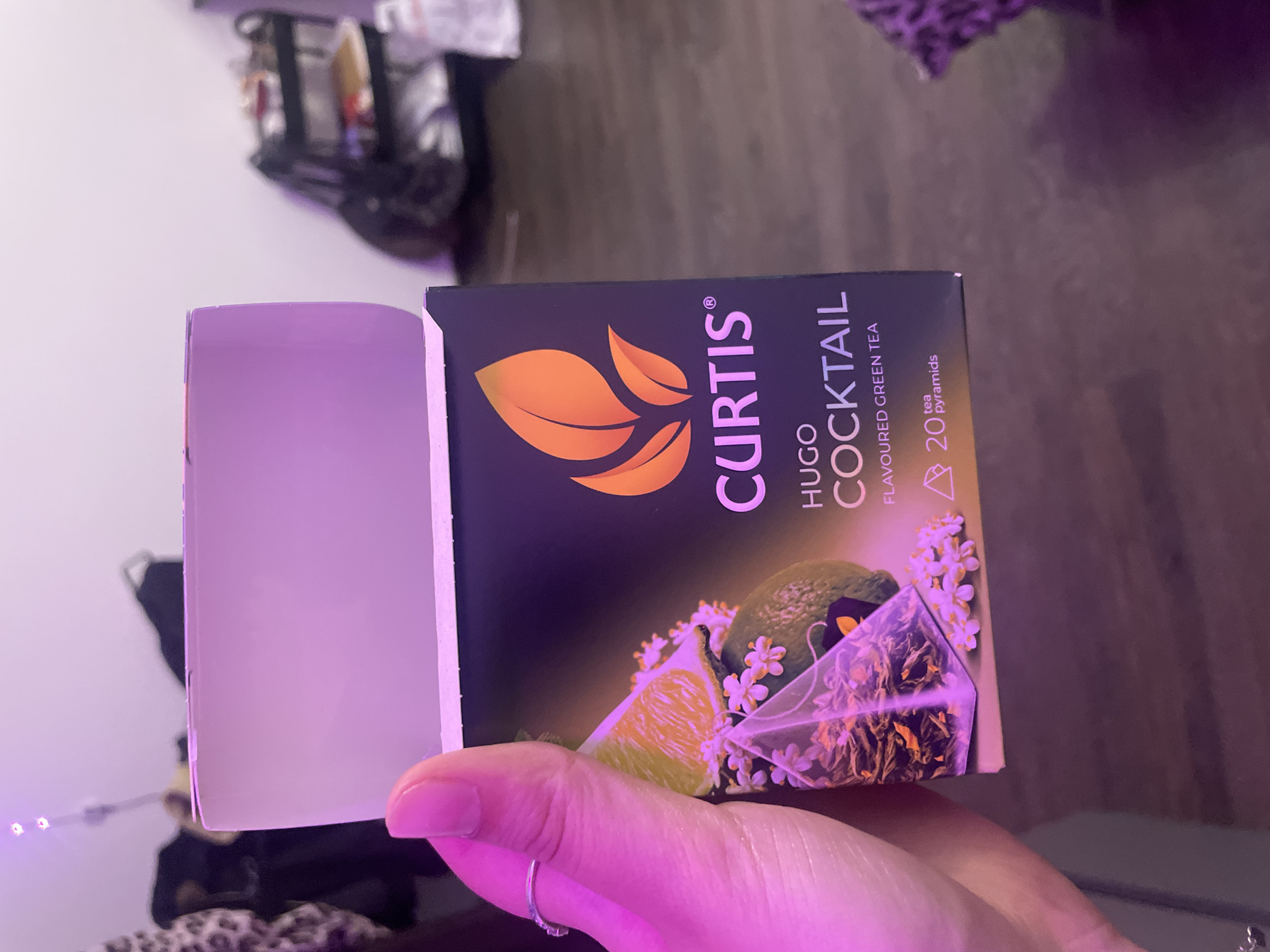 A hand, adorned with a silver ring on the index finger, holds a box of Curtis Hugo Cocktail flavored green tea. The box is dark purple with an orange leaf logo and text. The tea itself is visible through a transparent pyramid-shaped bag, filled with green tea leaves and some orange pieces, resting on a slice of lime and small white flowers. The background is blurred, showing a room with furniture and a purple light casting a glow. The image does not contain information about the city of Sinyavino, Russia.