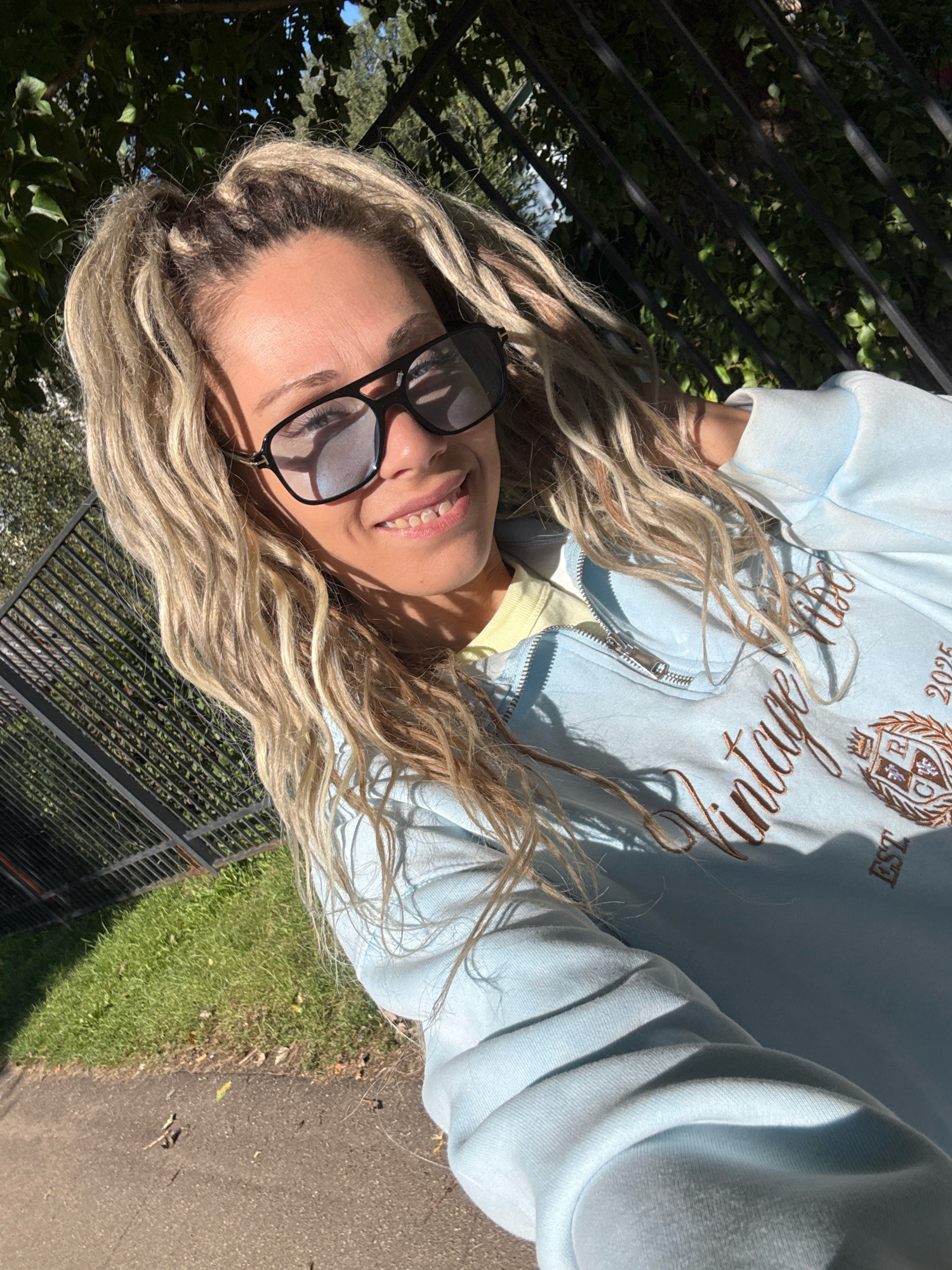 The image shows a woman with blonde dreadlocks, wearing large black sunglasses and a light blue zip-up hoodie. The hoodie has "Vintage Vibes" and "2025" embroidered in brown on the front, along with a crest. She appears to be taking a selfie outdoors, with a black metal fence and green foliage visible in the background. The ground in the foreground is a mix of pavement and grass. The lighting suggests it is a sunny day. The location is identified as Zhukovo, Russia.