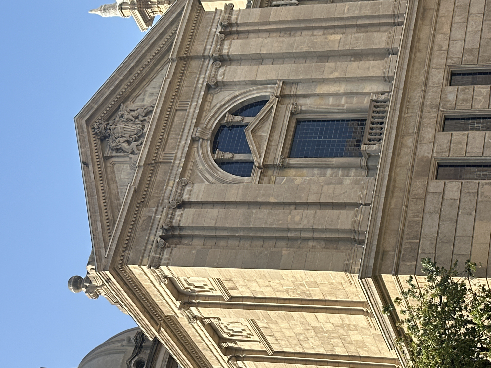 The image shows the facade of a grand stone building in Barcelona, Spain. The building has classical architectural elements, including columns, pediments, and decorative carvings. The facade is made of large, square stone blocks. The building is bathed in sunlight, casting shadows that highlight the architectural details. A bright blue sky is visible above. In the lower left corner, part of a green tree is visible.
The building depicted is Palau de la Generalitat de Catalunya, located in Plaça Sant Jaume in Barcelona. The architectural style is predominantly Gothic with Neo-Gothic additions. The specific section of the building shown is likely part of the more modern extensions, which incorporate Renaissance and Baroque influences, evident in the ornamentation and the pediment above the arched window with the coat of arms.
