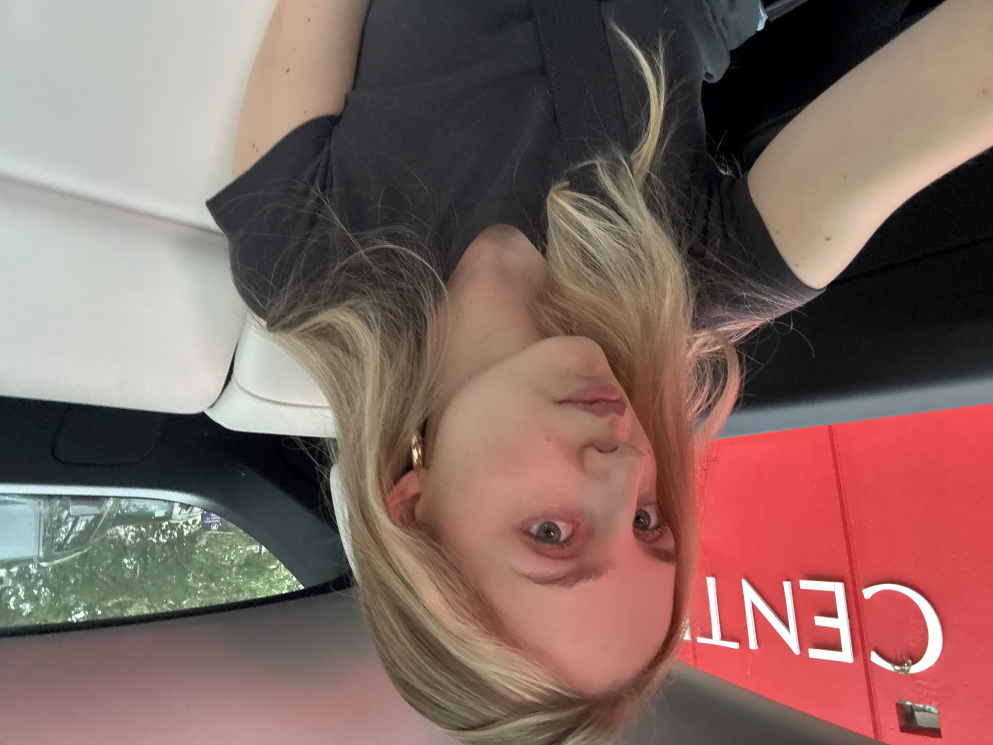 This is a vertical, medium shot of a young person with blonde hair and blue eyes, possibly taken from inside a vehicle. The person is wearing a dark t-shirt and a gold hoop earring. Their hair is long and falls around their face. The person's face is turned towards the camera, and they appear to be looking directly at it with a neutral expression.
In the background, to the right, there is a large red sign with white text that appears to say "CENT". The rest of the sign is cut off by the frame. The environment suggests an outdoor setting, possibly a commercial area or a public space, given the signage. The lighting suggests it could be daytime, though the specific time of day is not clear. The reflection in the window to the left shows trees and greenery, hinting at an exterior environment. The interior of the vehicle is visible, with white upholstery on the left side.