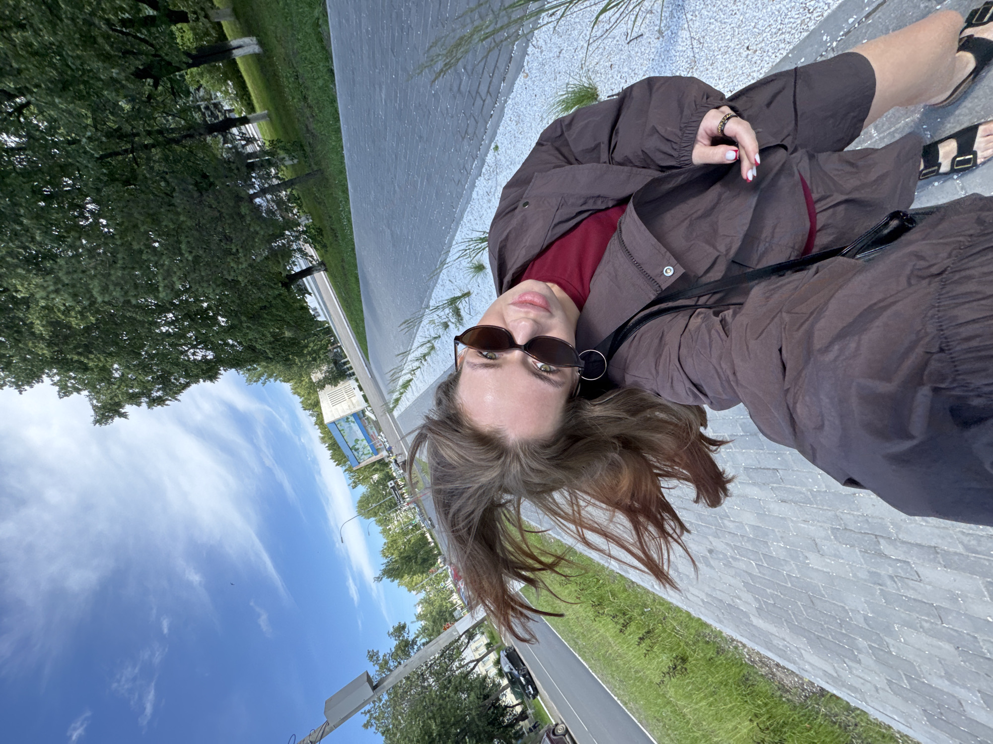 A young woman with long, brown, windswept hair is taking a selfie outdoors. She is wearing a dark brown jacket over a maroon shirt and brown shorts, with black sandals. She has on large, dark sunglasses and hoop earrings. Her right hand, with red-tipped fingernails and a gold ring, is visible. She appears to be walking on a paved sidewalk next to a grassy area and a road with a black car driving on it. In the background, there are trees, a street lamp, and a modern building with a colorful mural. The sky is blue with wispy white clouds. The scene is likely in the city of Chaikovsky, Russia, given the context.