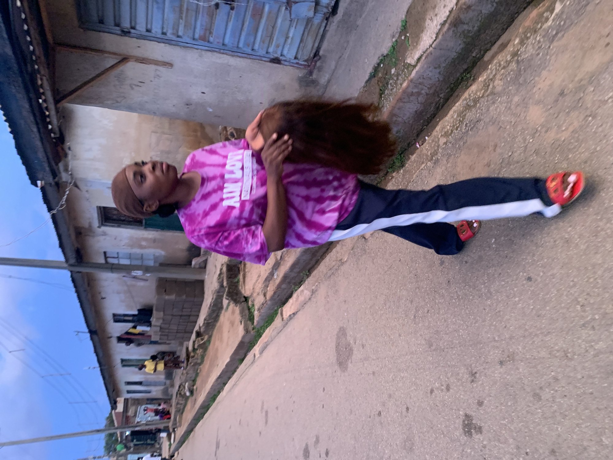 An outdoor, portrait-oriented photograph, taken in Jos, Nigeria, captures a young person holding a wig on a paved street. The subject, positioned in the midground, is likely female, wearing a vibrant pink and white tie-dye t-shirt, dark blue track pants with a white stripe down the side, and red open-toed sandals. She wears a light brown head covering and holds a long, flowing dark brown wig with both hands, allowing it to hang towards the ground. Her gaze is directed slightly upwards and to the left, with a neutral expression.
The street she stands on is paved, featuring a raised concrete curb and a narrow strip of sparse greenery to her right. The background showcases typical urban residential buildings under a clear, light blue sky, suggesting late afternoon or early evening. To the left, a multi-story building with a corrugated metal roof and worn, light-colored walls is visible, with clothes hanging out to dry from windows or balconies. Another individual in a yellow shirt is visible further down the street. Utility poles and overhead wires crisscross the sky in this section. To the right, another building with a closed metal shutter and weathered walls is partially visible.
On the subject's t-shirt, white text is visible, with "AIN" partially seen on the top line, and "LOVE" clearly legible below it, suggesting part of a slogan or brand. The scene depicts an everyday moment in a residential area of Jos, Nigeria.