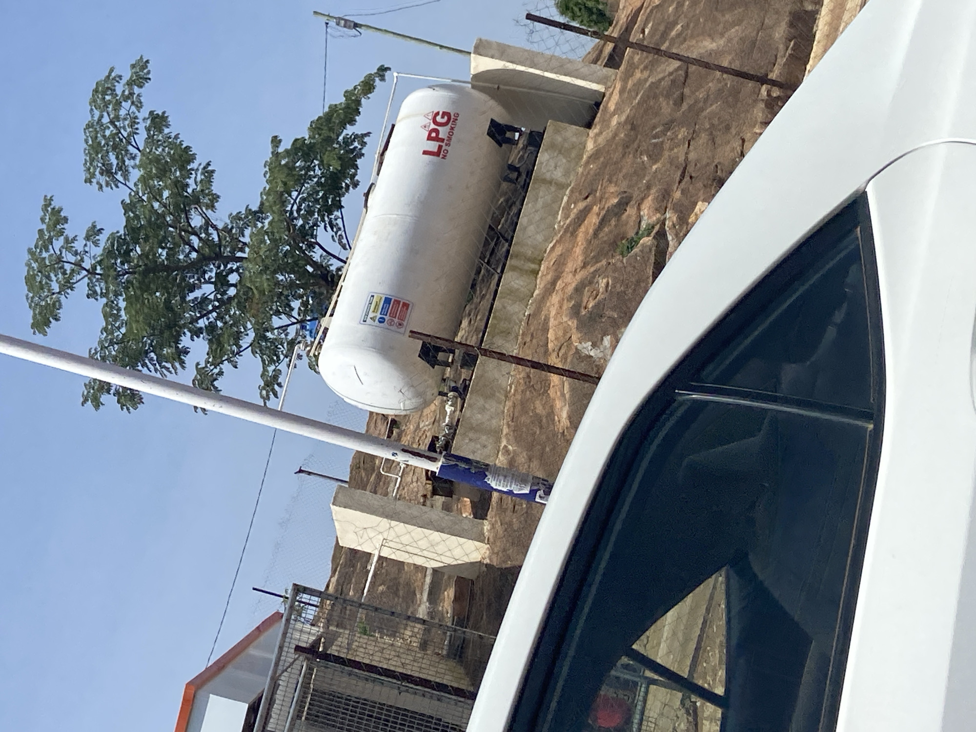 In the city of Jos, Nigeria, a white LPG gas tank is prominently displayed on a rocky, elevated terrain. The tank, cylindrical and horizontal, is white with "LPG NO SMOKING" written in red lettering. It rests on a concrete foundation secured by metal supports and enclosed by a chain-link fence. Adjacent to the tank, a tall white pole stands vertically, marked with a blue and white striped band near its base. In the foreground, a white car is partially visible, its tinted windows reflecting the surrounding environment. Behind the tank, a lush green tree with dense foliage provides a natural backdrop. To the left, a building with an orange roof and white walls is partly seen, along with a metal fence and a concrete structure. The sky above is clear and blue, indicating a sunny day.