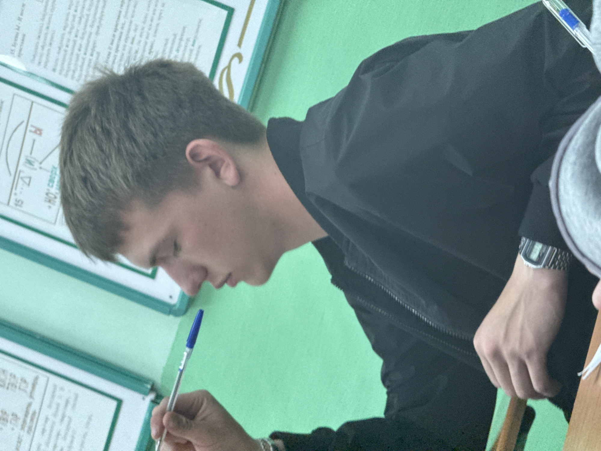 The image shows a young male, likely a student, seen in profile within an indoor setting, possibly a classroom. He has short brown hair and fair skin and is intently looking downwards, suggesting he is writing, reading, or drawing. He holds a blue pen in his right hand. A silver digital watch with a metal strap is visible on his left wrist. He is wearing a black jacket with a zipper. Part of a gray hooded garment is visible on the right side of the frame, possibly belonging to another person sitting nearby.
The background consists of a light green wall decorated with several framed documents or posters, each with a white background and a green border, reinforcing the classroom or study environment. Visible Cyrillic text on the top left poster includes `15°`, `Δ (М) — Ы`, and `НО сверху`, indicating content that could be related to mathematics, geometry, or a scientific subject. A wooden surface, possibly part of a desk or chair, is also visible on the right. The young man appears focused and calm, showing no strong emotions other than concentration. The lighting is even, and the image is slightly tilted.