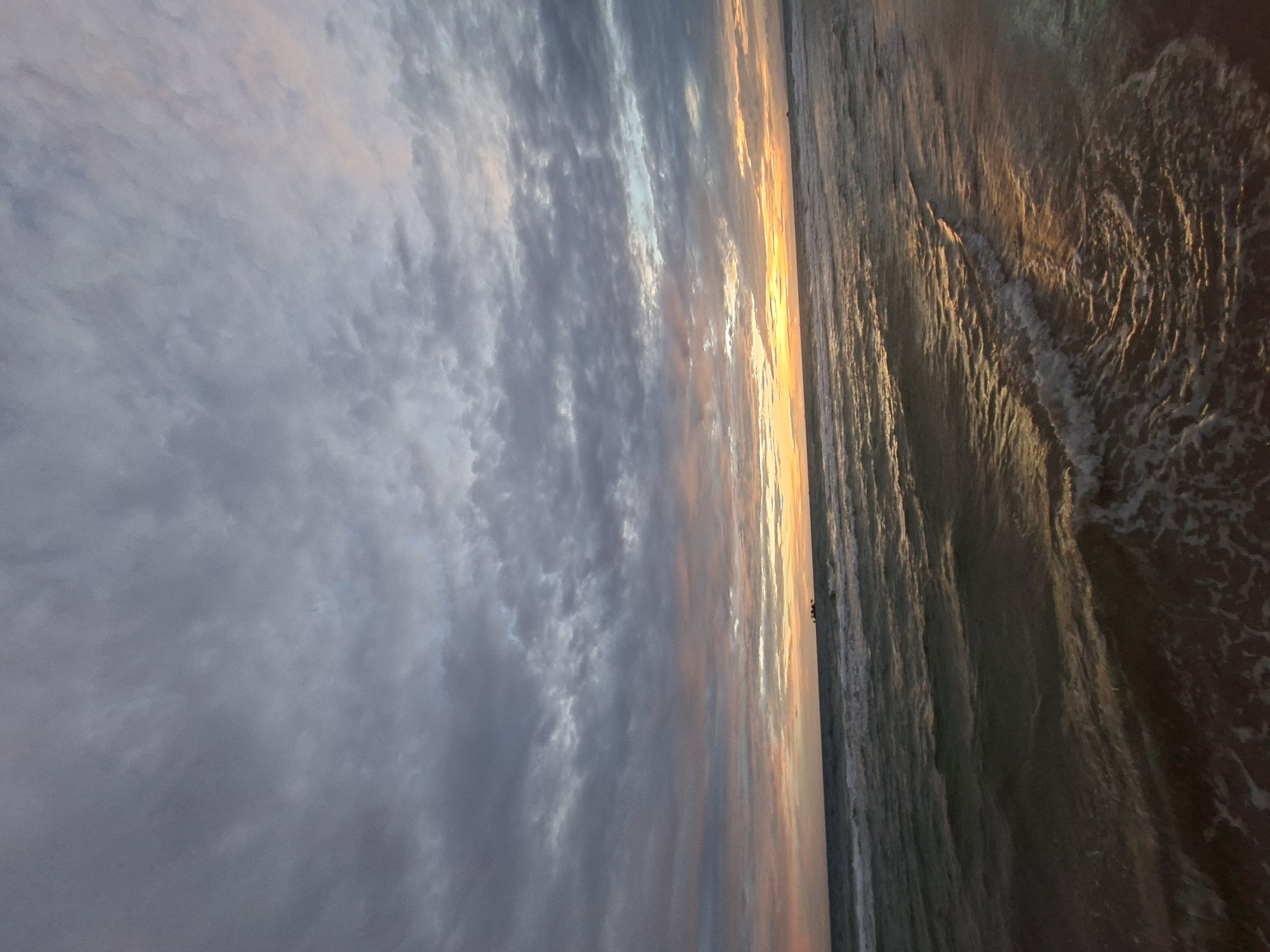 An expansive view of a beach at sunset shows the ocean meeting the shore under a cloudy sky. The water is a deep teal with small waves and white foam cresting. The sky above is filled with gray, white, and pink clouds, with the setting sun creating a warm, orange glow on the horizon. A small dark object, possibly a boat or debris, is visible on the water in the distance. The sand is not clearly visible, but the edge of the water suggests a sandy beach. The location is identified as Phú Quốc, Vietnam.