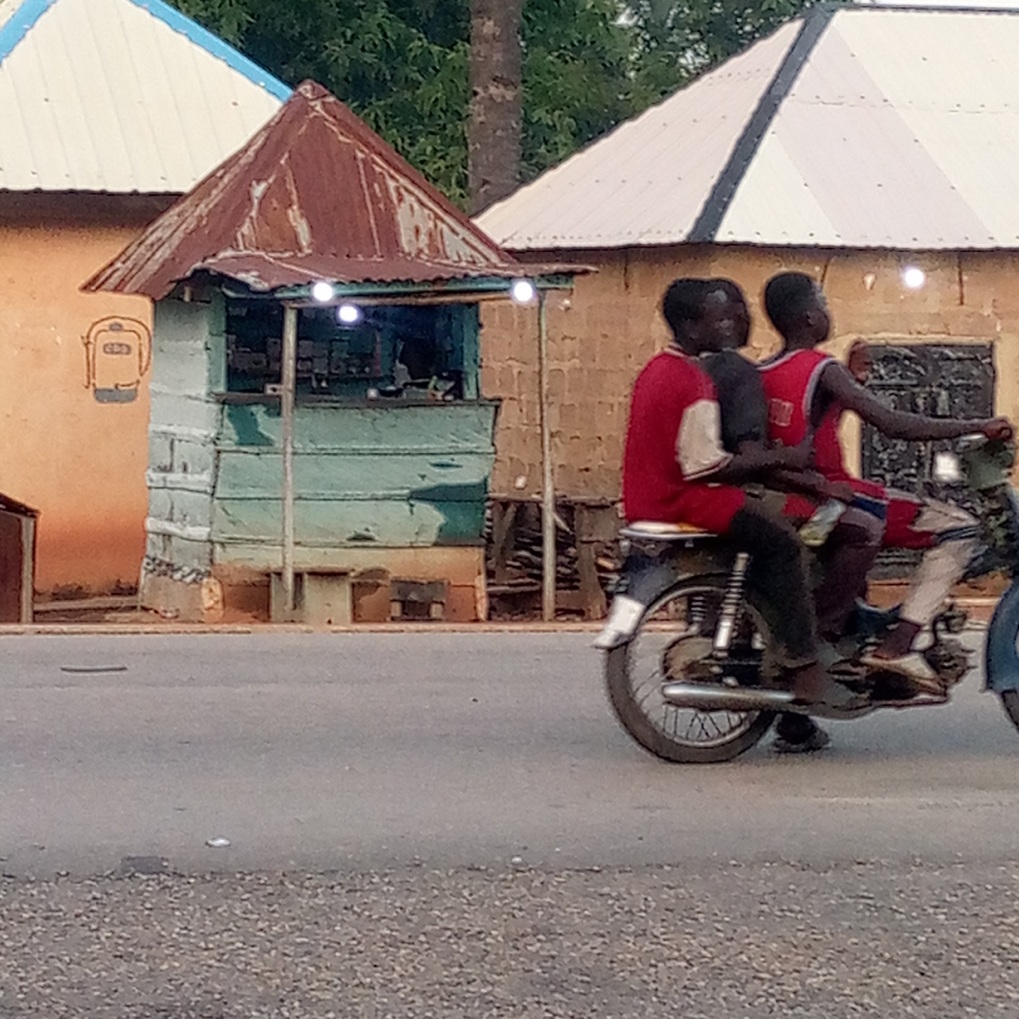 The image depicts a street scene in Mararaba, Nigeria, likely captured during the late afternoon or early evening given the soft lighting and activated lights. In the midground, three individuals, appearing to be young men or boys, are riding a dark-colored motorcycle. The person in front and the one directly behind him wear red tops, while the third rider at the very back is partially obscured.
To the left, a small, rustic kiosk or stall stands prominently, featuring a heavily rusted, pointed corrugated metal roof and faded light-green or blue painted walls. Two small, bright white lights are visible within or mounted on the front of the kiosk, suggesting it is either operational or providing illumination. Behind this kiosk, a building with an orange-tan wall displays a distinctive hand-painted graphic that resembles a backpack or satchel, with some faint, indistinct lettering within it. Further back, parts of other buildings with white corrugated roofs and green foliage are visible. The foreground shows a paved road with scattered gravel along the shoulder. No clearly legible text is visible other than the indistinct letters on the painted graphic.
