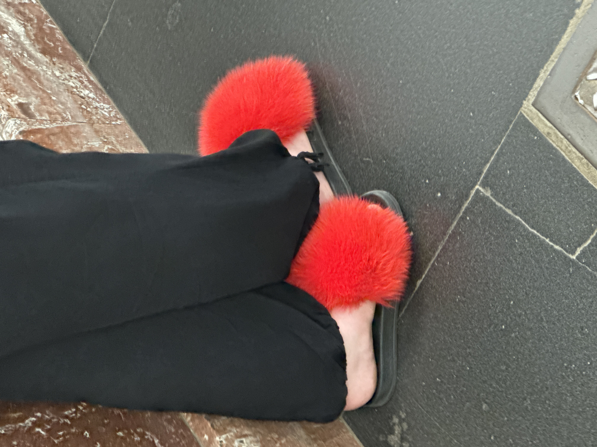 The image displays the lower body of a person, from mid-calf level downwards, situated in an indoor environment identified as a location in Saint Petersburg, Russia. The person is wearing loose-fitting black pants that obscure the upper portion of their feet. On their feet are bright red, heavily furred slides with visible black soles, revealing the bare heels and parts of the arches of the feet, which have a light skin tone. The person is positioned adjacent to a reddish-brown wall or pillar on the left side of the frame, which exhibits prominent white, streaky patterns consistent with marble. The floor is constructed from dark grey or black textured tiles, delineated by lighter grout lines, and shows scattered small, light-colored specks, indicative of dust or debris. In the bottom right quadrant of the image, a partial view of a square metal plate or access panel with a lighter border is embedded within the tiled floor.