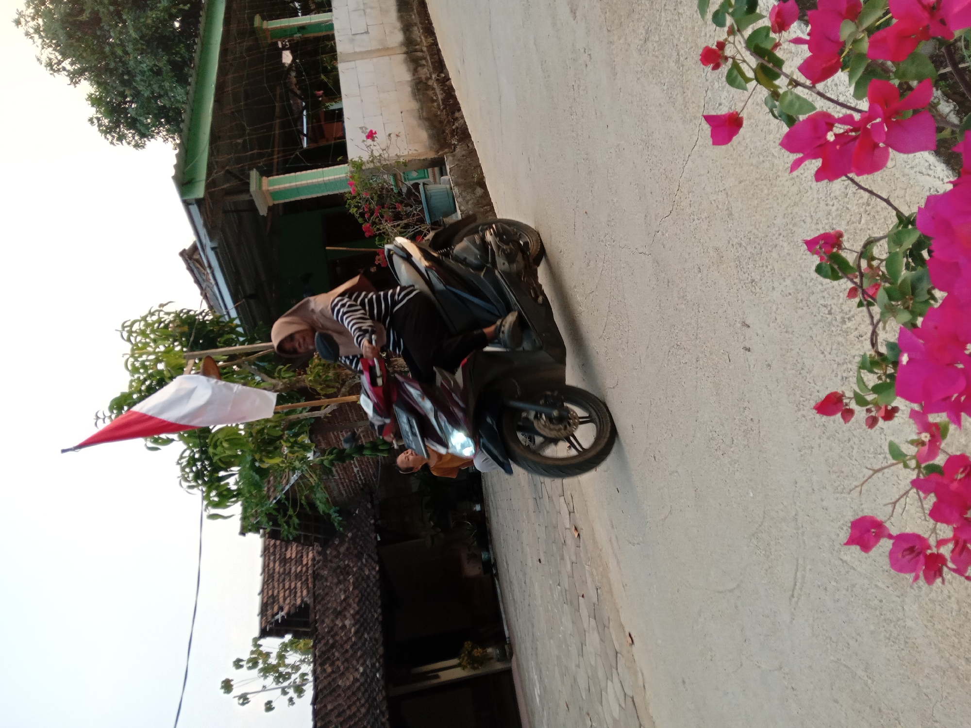 A woman wearing a hijab and a striped shirt rides a scooter on a paved road in what appears to be a rural or suburban area. An Indonesian flag is planted behind her on a pole. In the background, there are houses with tiled roofs, trees, and a white wall. Pink flowers are visible in the foreground. Another person, possibly a child, stands in the background near a house. The lighting suggests it is late afternoon or early morning.