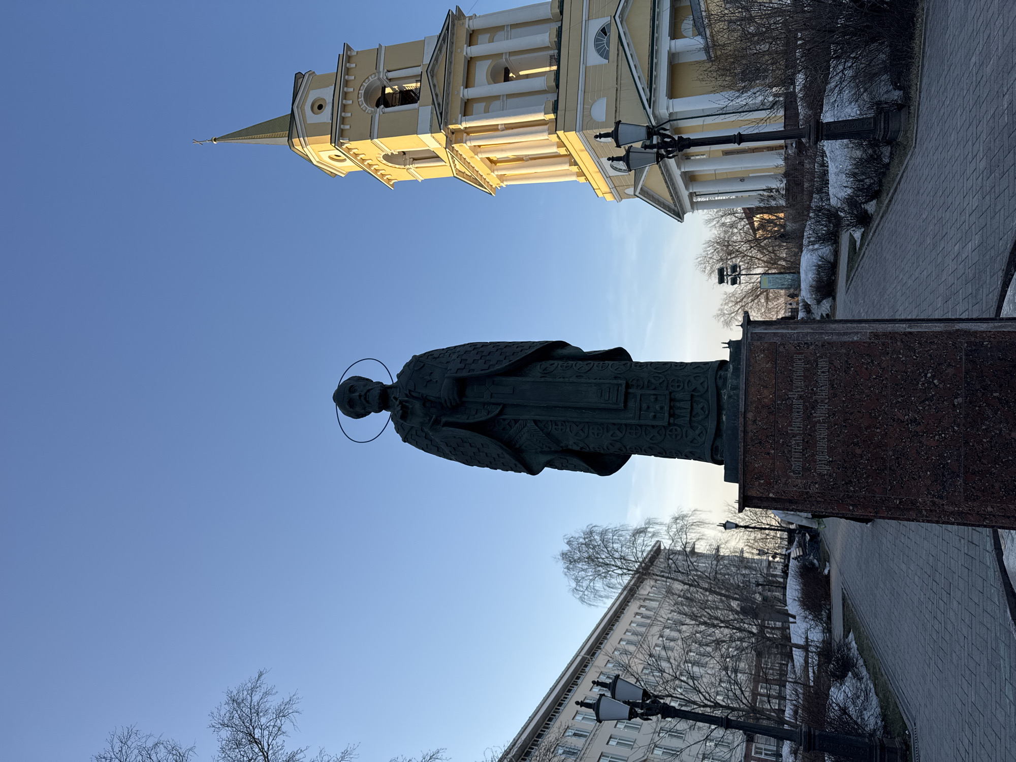 The image shows a monument of Saint Nicholas the Wonderworker in Perm, Russia. The statue is positioned in front of a yellow building with a tall bell tower and an adjacent modern building. The statue faces forward with a halo above its head. The scene is captured during the daytime under a clear sky. The ground is covered with pavement, with bare trees and light posts visible.
