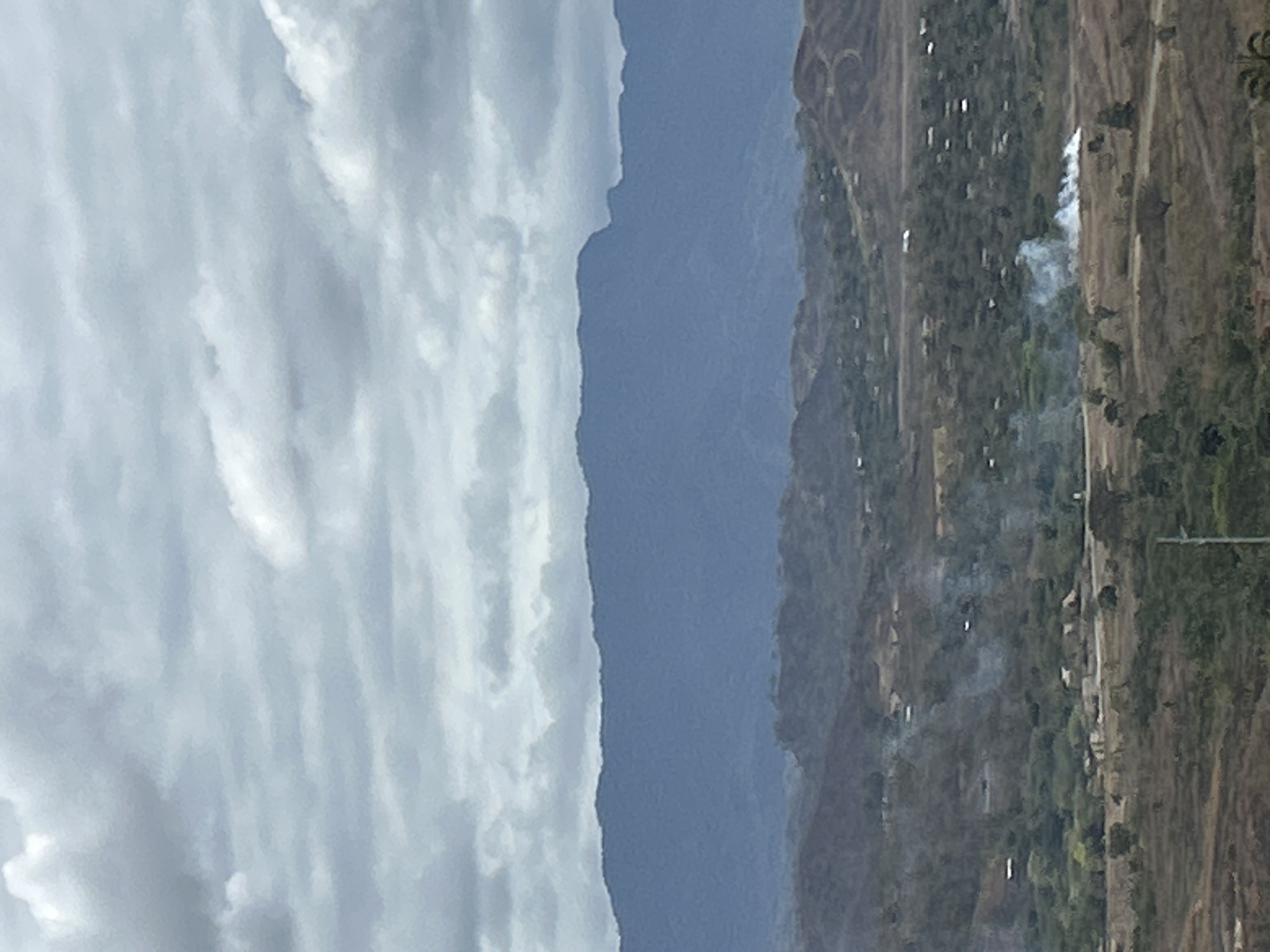 This image depicts a wide, hazy landscape view from an elevated position overlooking a rural or semi-rural area that is likely on the outskirts of Port Moresby, Papua New Guinea. The sky is overcast with heavy, gray clouds. In the middle ground, there are rolling hills covered with sparse vegetation and dotted with small settlements, indicated by white roofs of buildings. There is evidence of smoke plumes rising from a few areas within these settlements, suggesting fires or agricultural burning. The foreground consists of dry, brownish vegetation. In the far background, a large, dark blue mountain range forms the horizon, its details softened by distance and atmospheric haze. The overall impression is one of a vast, somewhat arid environment under a cloudy sky.