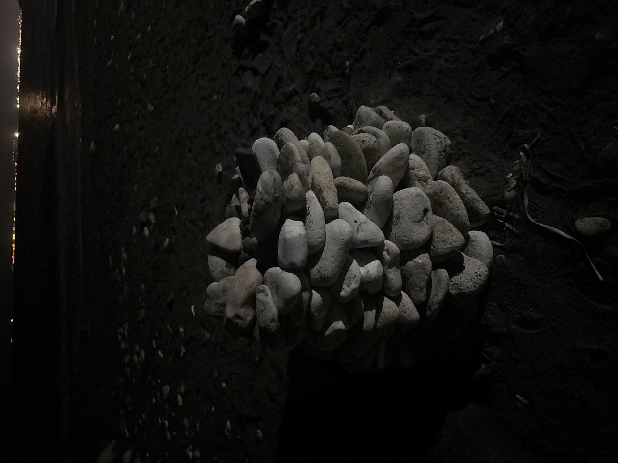 A nighttime, eye-level, medium shot captures a stacked pile of smooth, light-colored stones on a dark sandy beach in Agios Tychona Community, Cyprus. The stones are arranged in a roughly conical shape, with each layer slightly offset from the one below, creating a textured surface. The sand is dark and appears damp, with subtle ripples and a few scattered smaller stones. In the background, the calm sea is visible, reflecting the distant lights of what appears to be a coastline. The sky is dark, indicating it is nighttime. No people are present in the frame, and no actions or interactions are depicted. The overall scene is still and quiet.