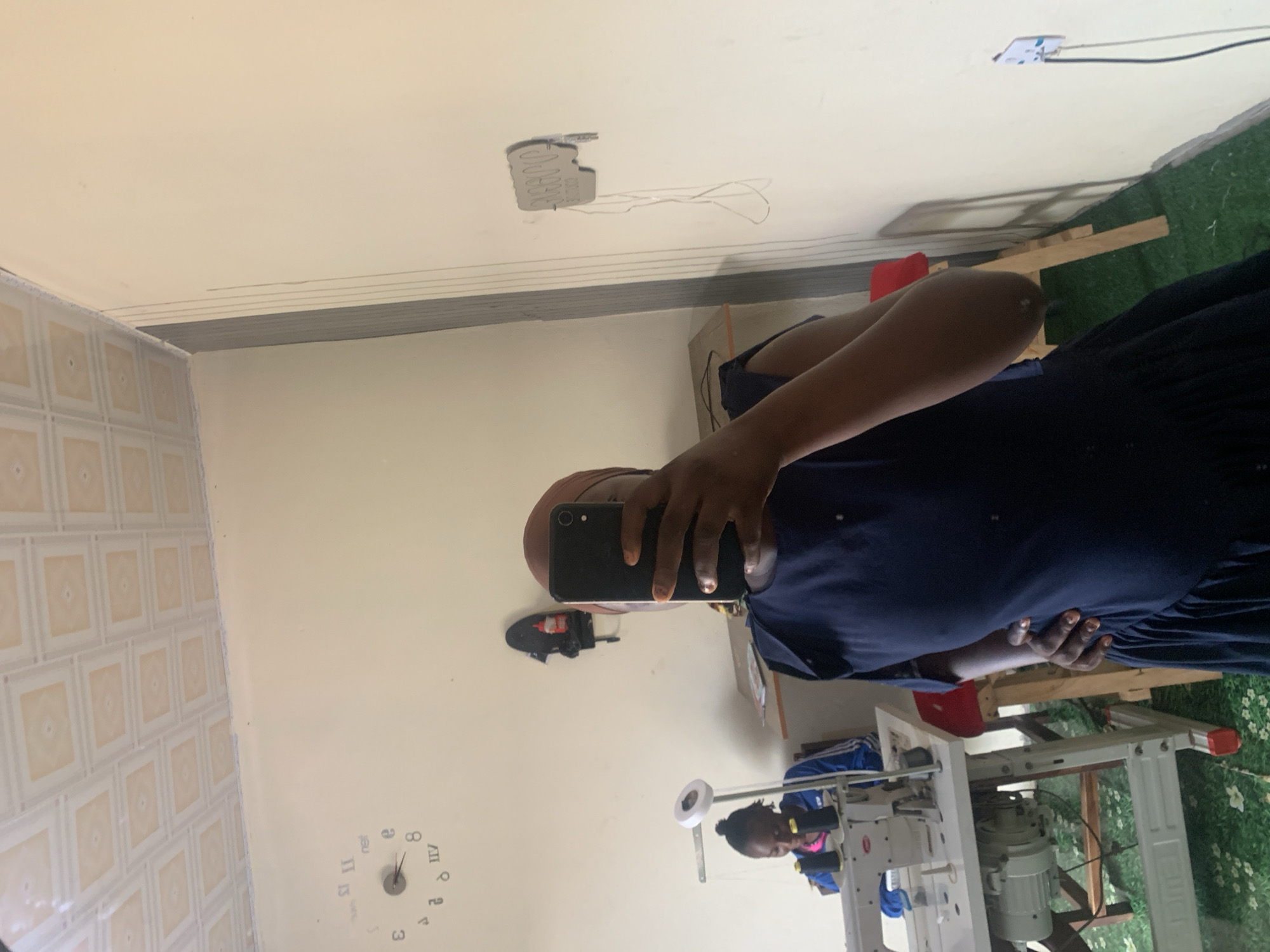 The image depicts a reflection in a mirror in what appears to be a workshop or tailoring business located in Jauro Yinu, Nigeria. In the foreground, a person is taking a selfie, holding a smartphone in their right hand and partially obscuring their face. They are wearing a dark blue dress and a brown headscarf. To the left, reflected in the mirror, is a young woman seated at a white industrial sewing machine. She is wearing a blue jacket with white stripes and a pink top. The workshop has light-colored walls and a patterned ceiling. A clock with Roman numerals is visible on the wall, and a power outlet with a black cable is also present. The floor is covered in what looks like green artificial turf with a floral pattern. The overall impression is of a functional workspace.