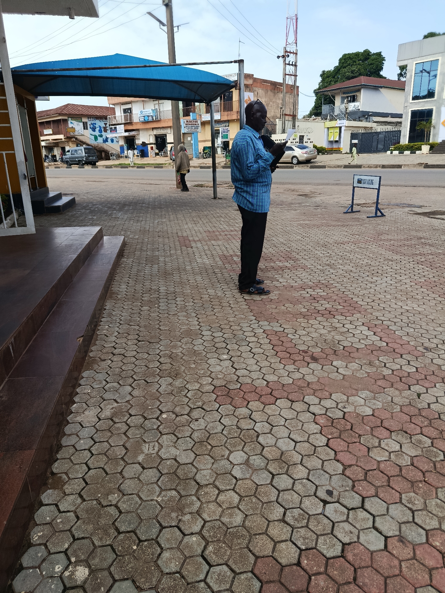 A man wearing a blue plaid shirt and black trousers stands on a paved sidewalk, looking down at a digital tablet he holds in his hands. He is wearing sandals. Behind him, a blue awning covers a sidewalk leading to several storefronts. A grey SUV is parked in front of one of the buildings. Further down the street, a tall radio tower is visible against a cloudy sky. The sidewalk is paved with hexagonal interlocking stones in red and grey. In the background, there are buildings and trees, and a sign with "CAR PARKING" written on it. The scene appears to be in a town or city, with shops and street infrastructure present. The lighting suggests it is daytime, possibly overcast.