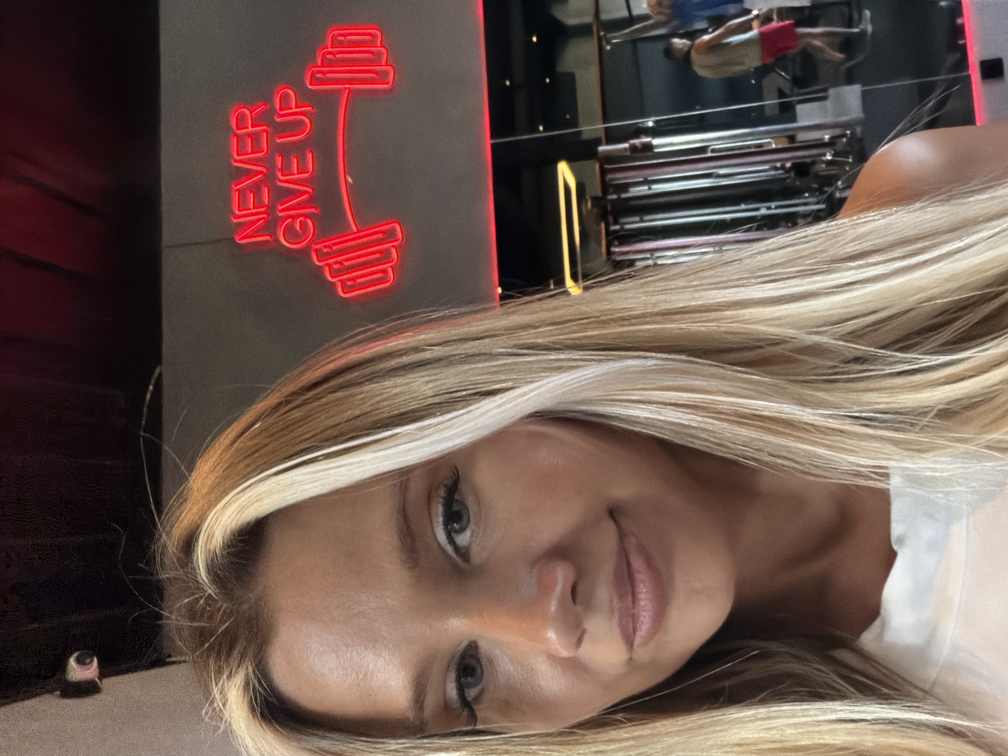 A young woman with blonde hair streaks and blue eyes takes a selfie in what appears to be a gym. She is wearing a white top. Behind her, a neon sign reads "NEVER GIVE UP" in red letters, above a red outline of a barbell. In the background, visible in a mirrored wall, are gym equipment and another person wearing a red pair of shorts and a grey tank top. The overall setting suggests a fitness establishment. The location is the city of North Kuta, Indonesia.