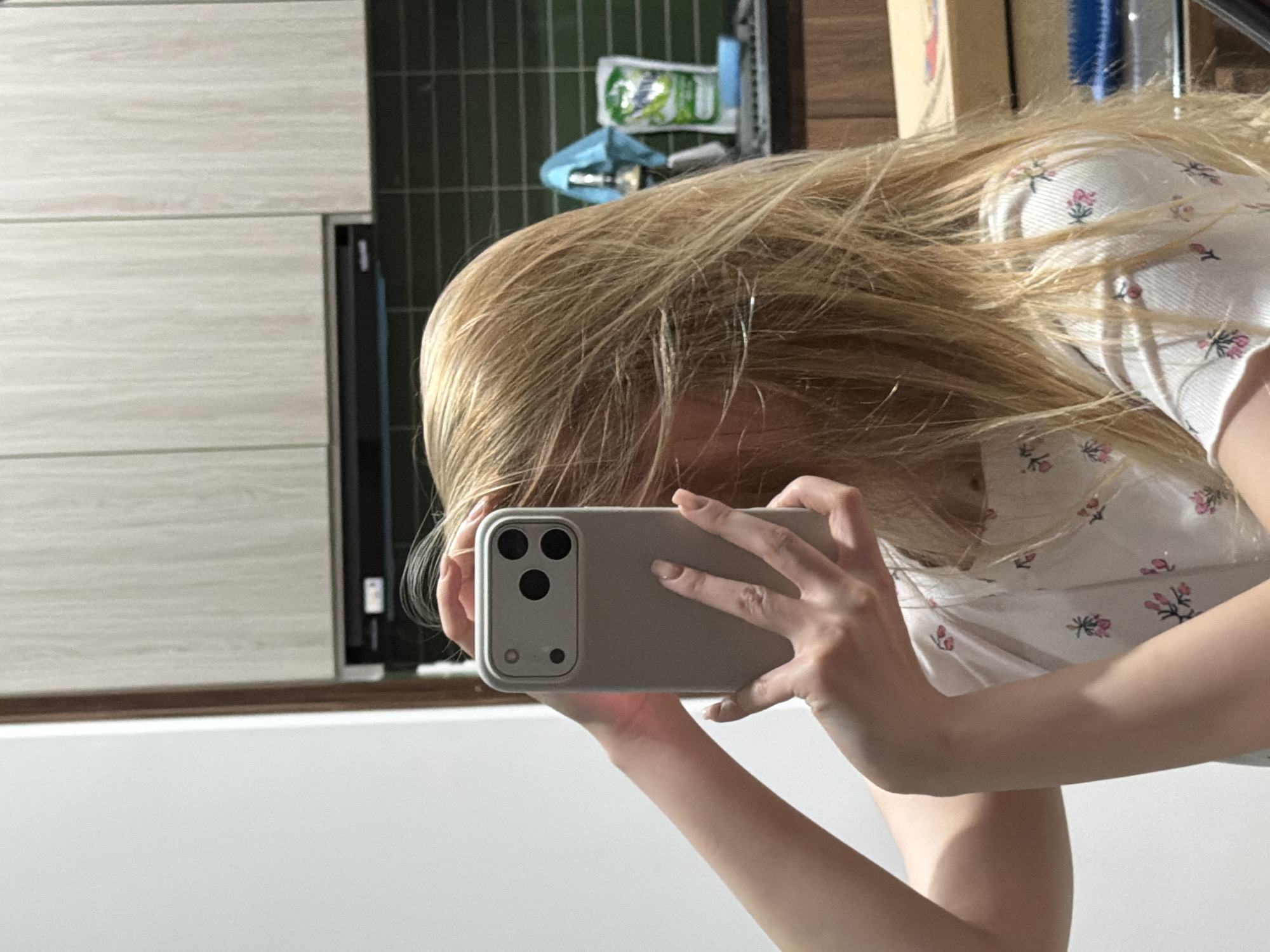 A person is holding a phone to take a selfie. The person has blonde hair and is wearing a white shirt with a floral pattern. The phone has a gray case and multiple camera lenses. The background includes a kitchen with white cabinets and green tiles. The location is not discernible in this image.