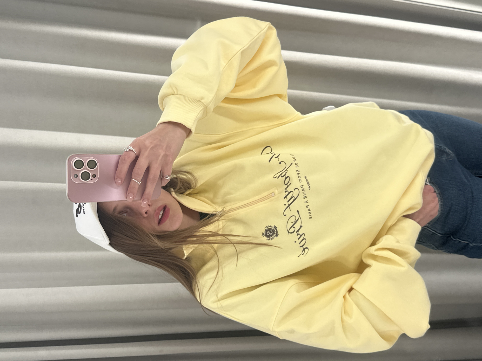 A young woman with long, brown hair is taking a selfie in a room with light gray curtains. She is wearing a white baseball cap, a light yellow quarter-zip sweatshirt, and dark blue jeans. The sweatshirt has a dark emblem and text on the front. She is holding a pink smartphone in her right hand, with her left hand resting in her jeans pocket. The scene appears to be indoors, possibly a fitting room or bedroom, with no discernible Moscow landmarks or indicators of location.