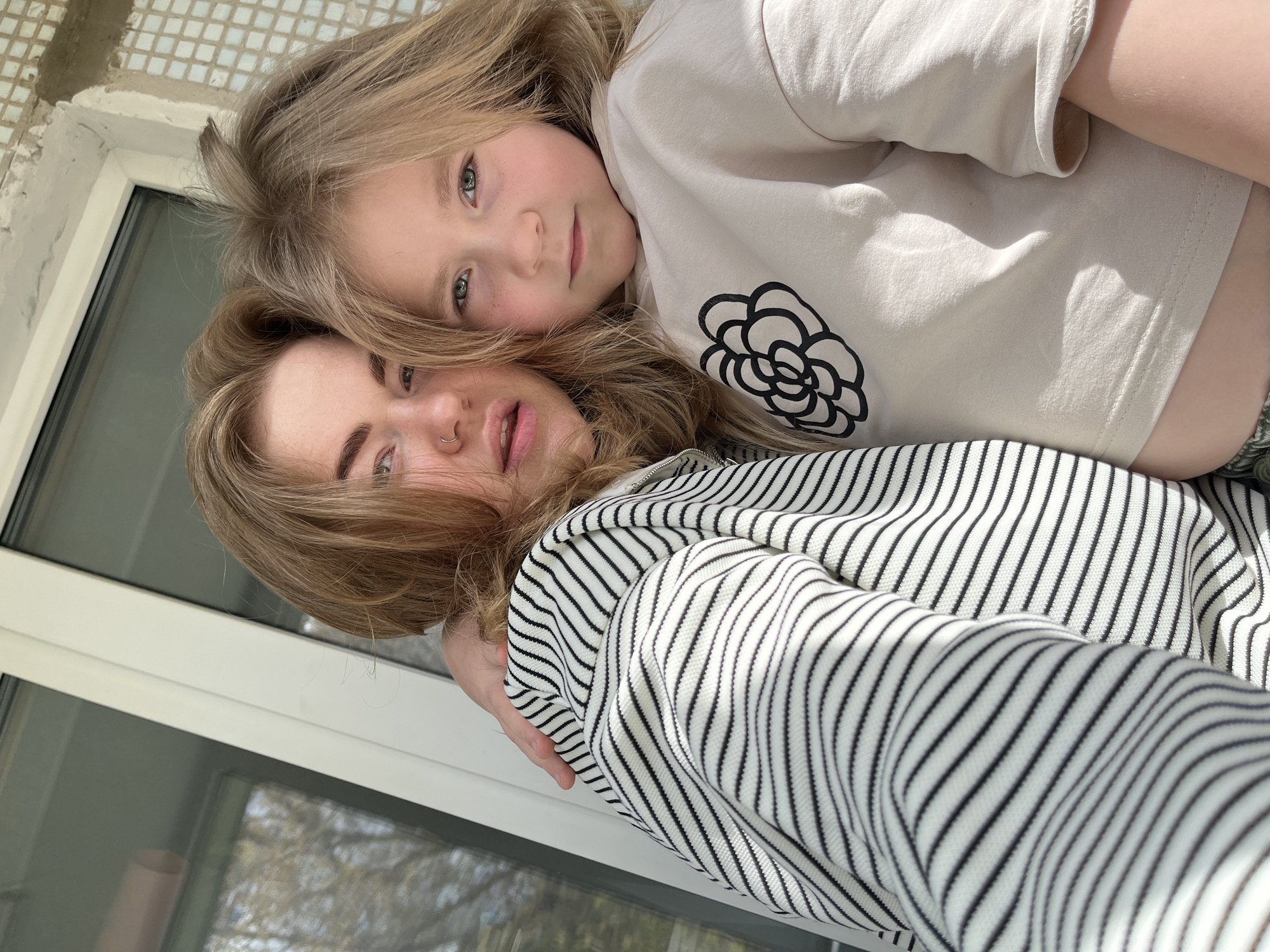 A medium-shot, tilted, eye-level photograph shows a blond-haired woman and a blond-haired girl embracing indoors, possibly in an apartment. The woman, wearing a black and white striped shirt, is on the bottom, and the girl, with fair skin and freckles, is on top, wearing a light tan shirt with a black flower design. The environment includes a white window frame, suggesting a window with a view outside. The scene does not provide enough information to confirm the location in the city of Chaikovsky, Russia.